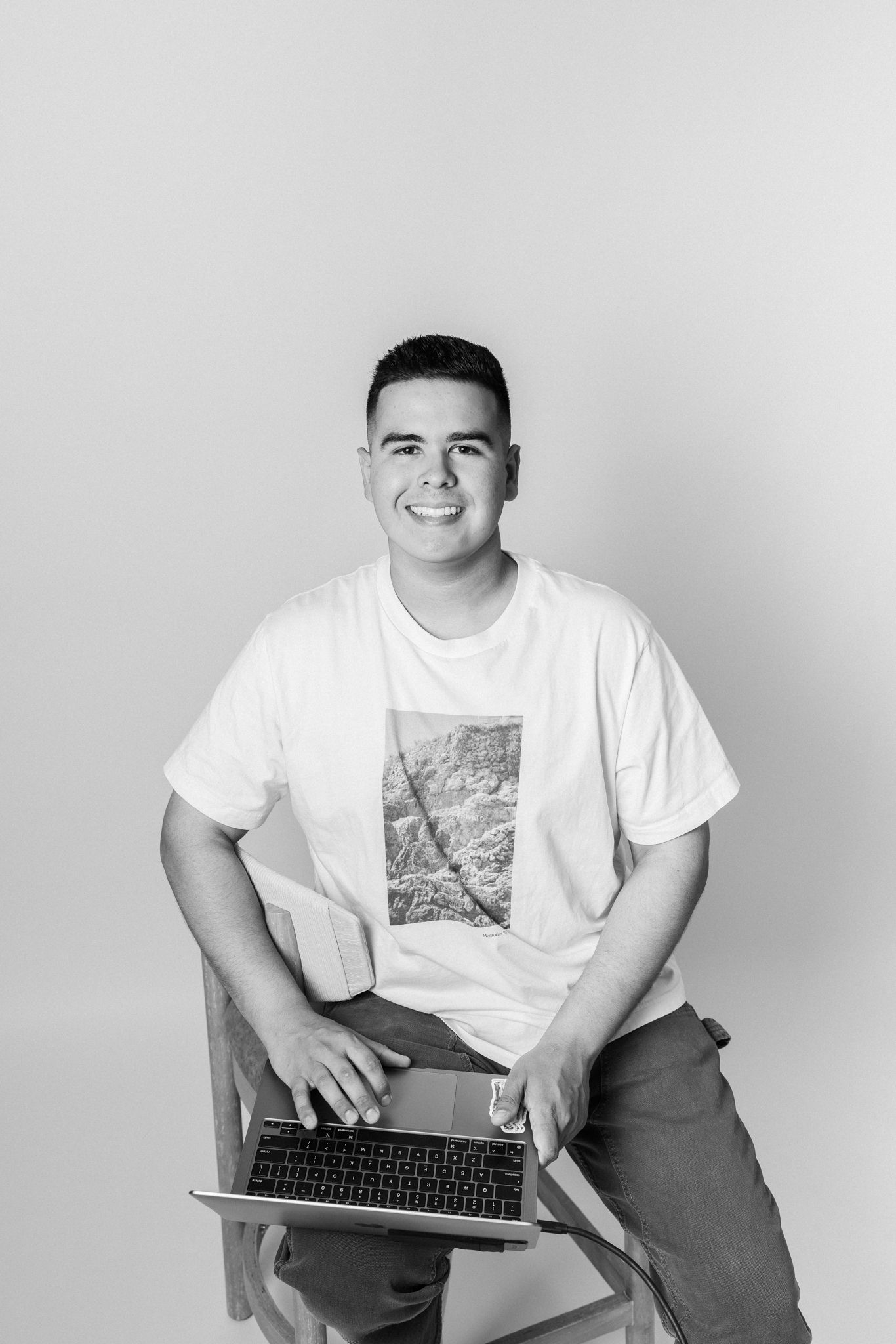 Young man smiling while sitting with a laptop in a black and white studio portrait.