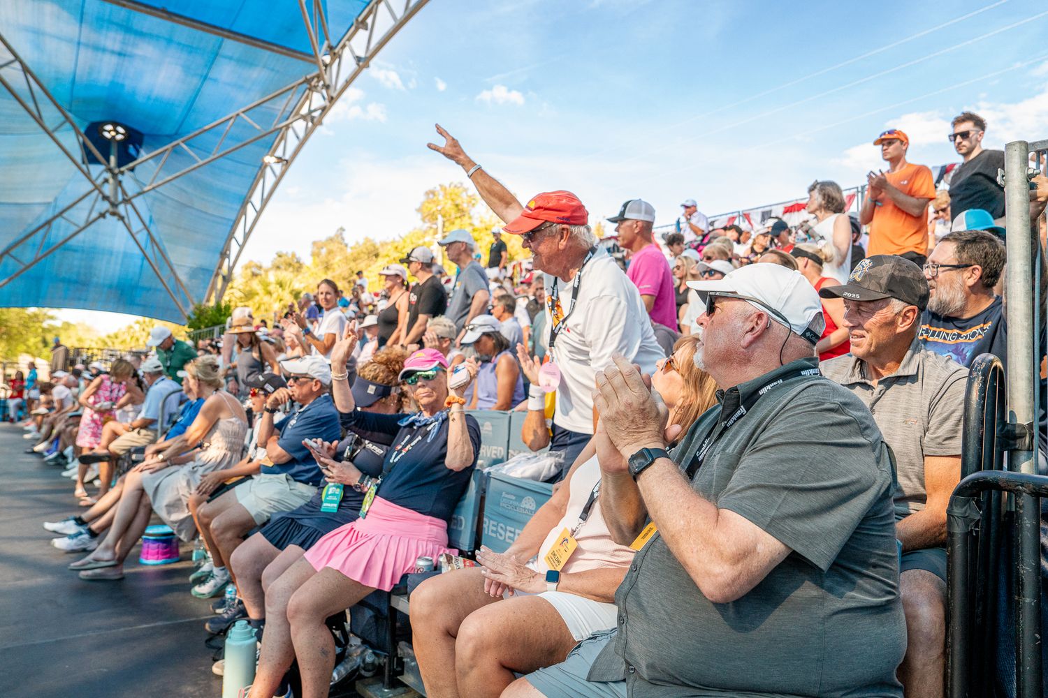 Large crowd of spectators enjoying an outdoor sports event under a blue canopy on a sunny day.