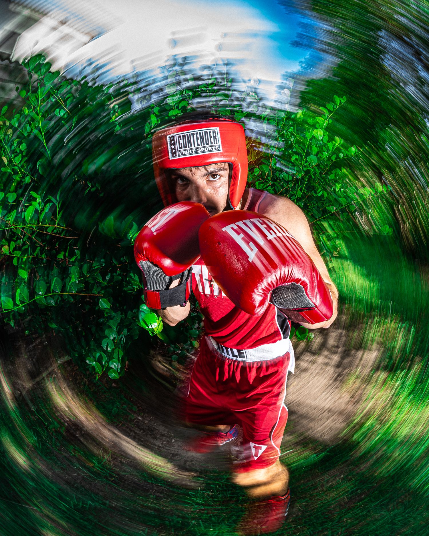 A young boxer in red Everlast gloves and Contender headgear throws a punch outdoors with a dramatic fisheye lens effect.