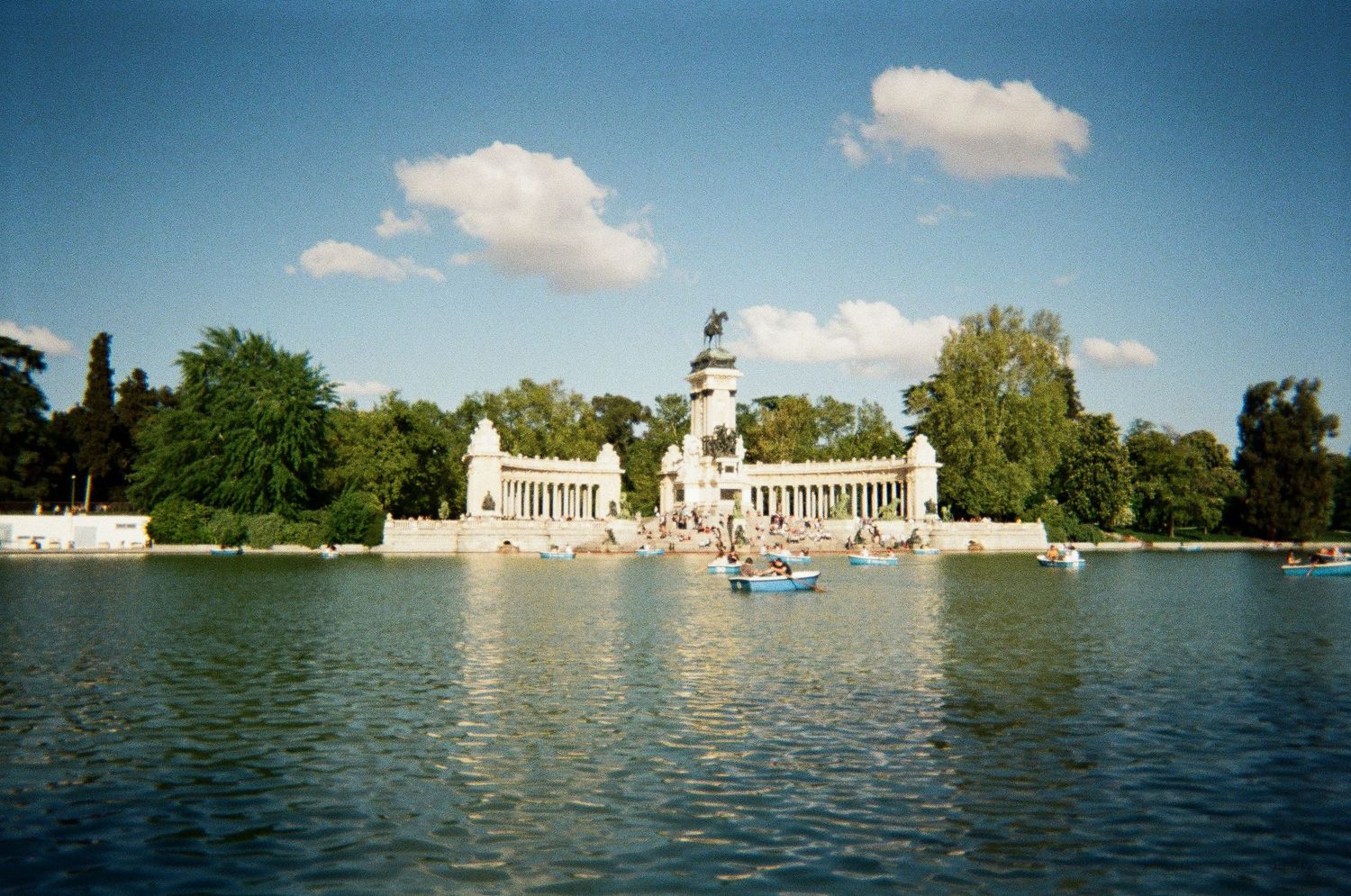 Scenic view of Retiro Park lake in Madrid with rowboats and monument under a blue sky.
