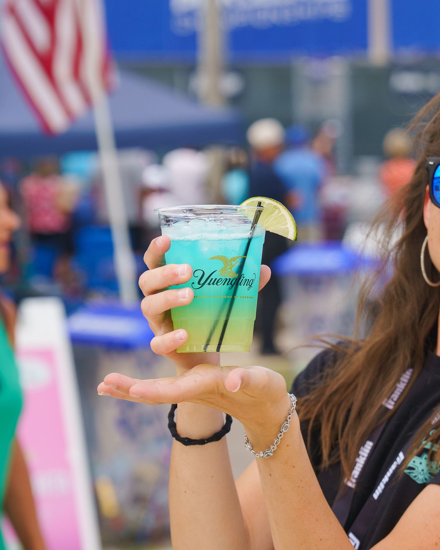 Woman holds a blue Yuengling cocktail with lime garnish at an outdoor sports event.
