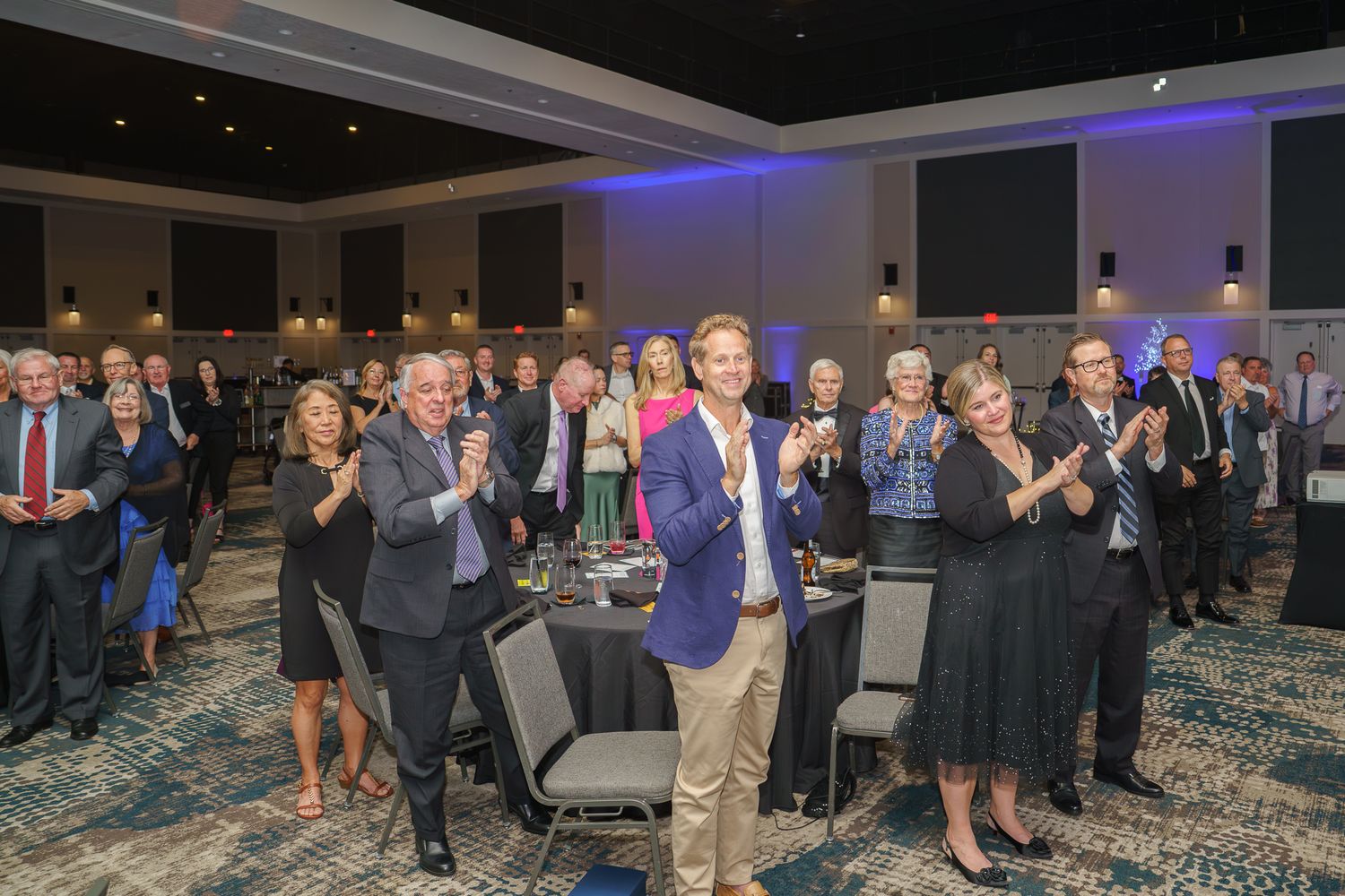 Group of well-dressed professionals applauding at an elegant blue-lit banquet hall event.