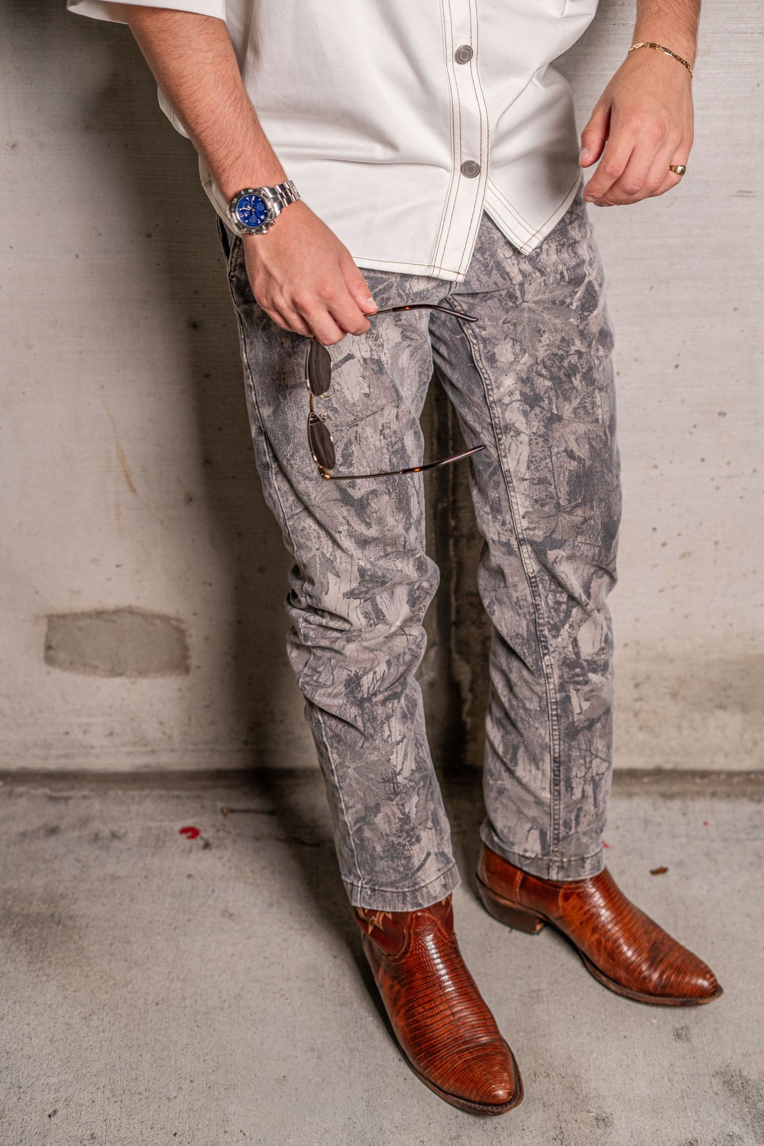 Man wearing gray camo cargo pants, brown cowboy boots, and a blue watch poses against a concrete wall.