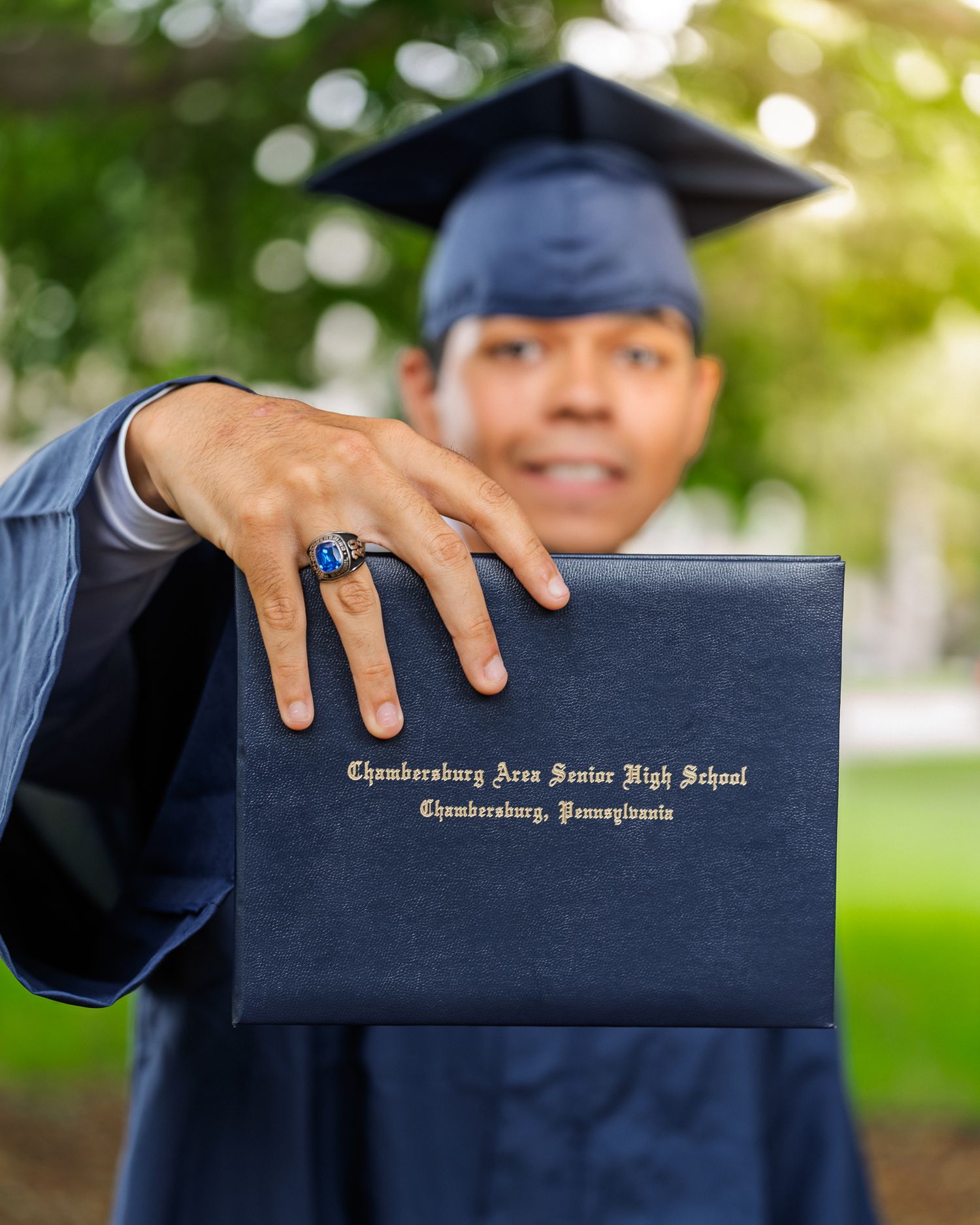 Graduate in cap and gown holds Chambersburg Area Senior High School diploma toward camera with class ring visible.