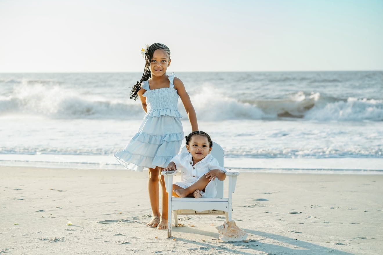 Two children pose on a sandy beach, girl in blue dress standing beside toddler boy seated in white chair with ocean waves behind.