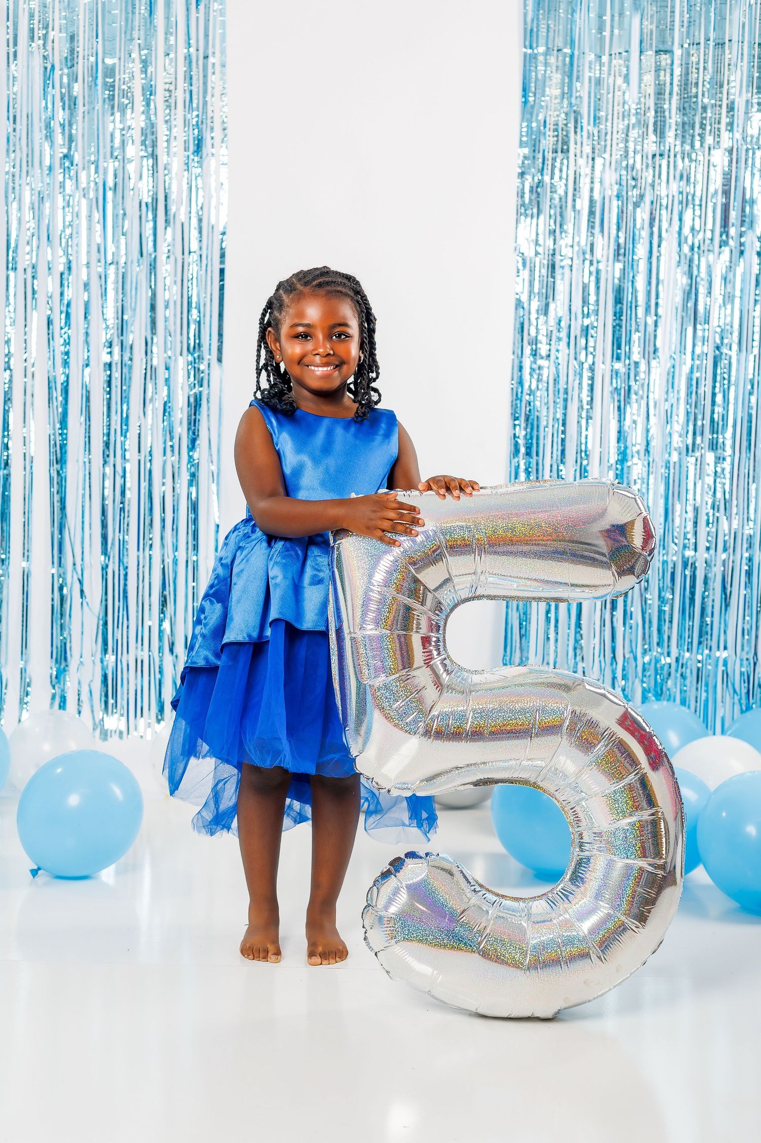 Young girl in blue dress stands holding large silver number 5 balloon surrounded by blue balloons.