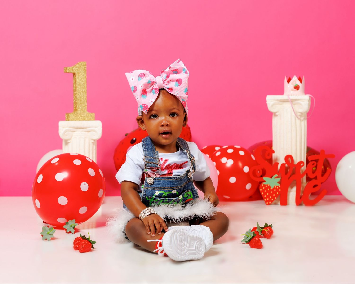 Baby girl with bow headband sits among red polka dot balloons and strawberry decorations for first birthday.