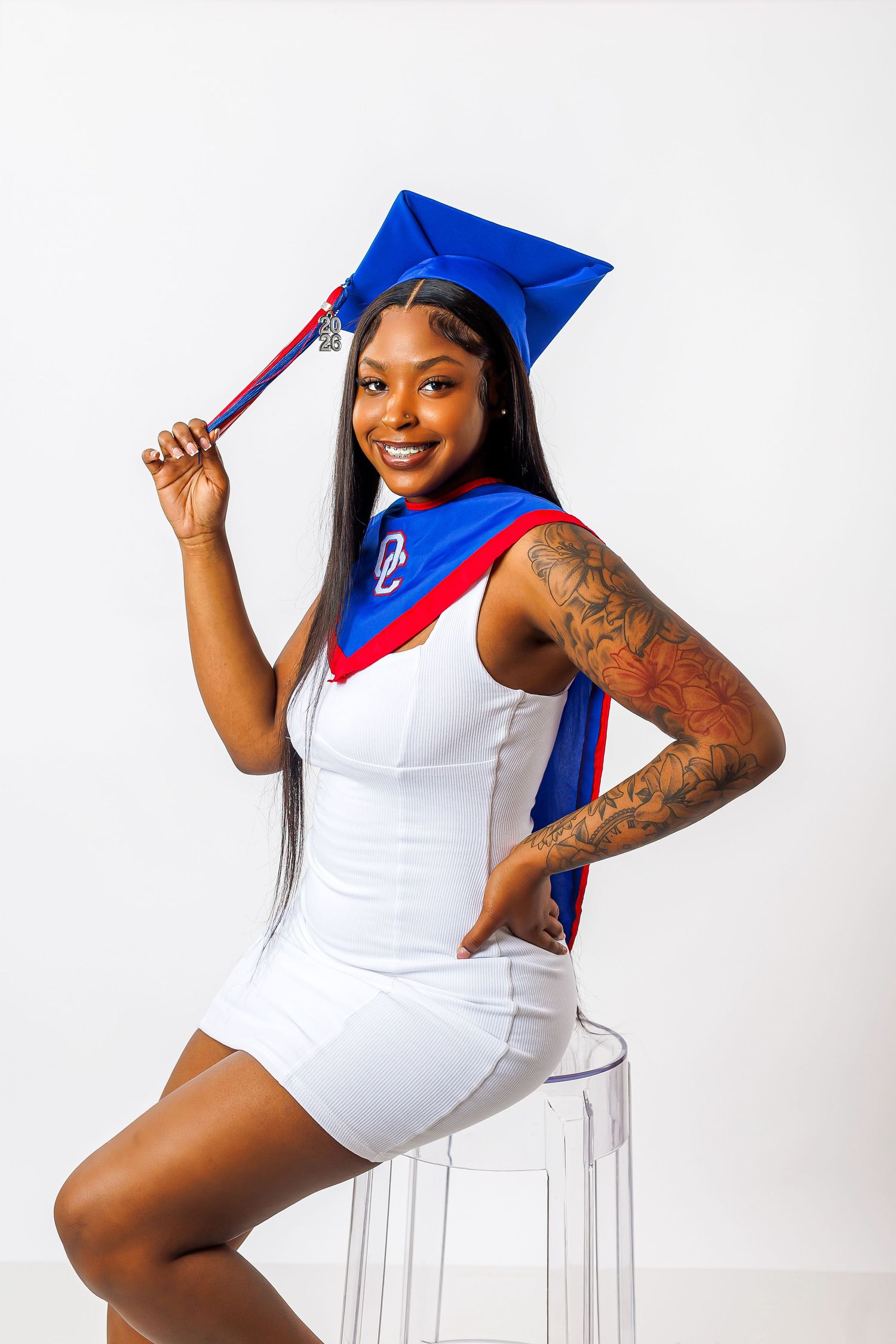 Tattooed graduate posing in blue cap and stole wearing white dress on a clear acrylic stool.