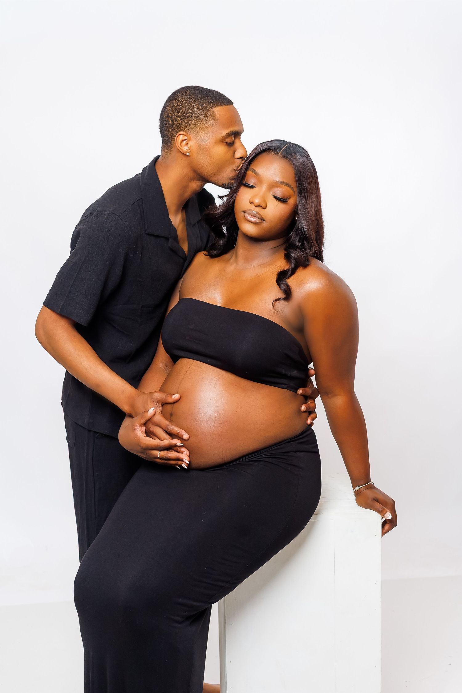 A man in black kisses a pregnant woman wearing a black bandeau top in a studio maternity shoot.