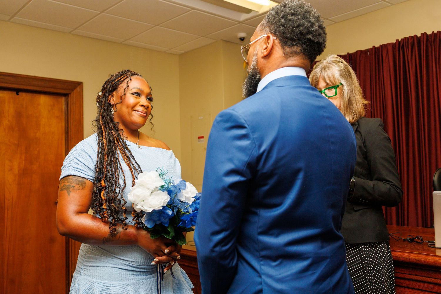 Bride holding blue bouquet exchanges vows with groom in blue suit at courthouse wedding.