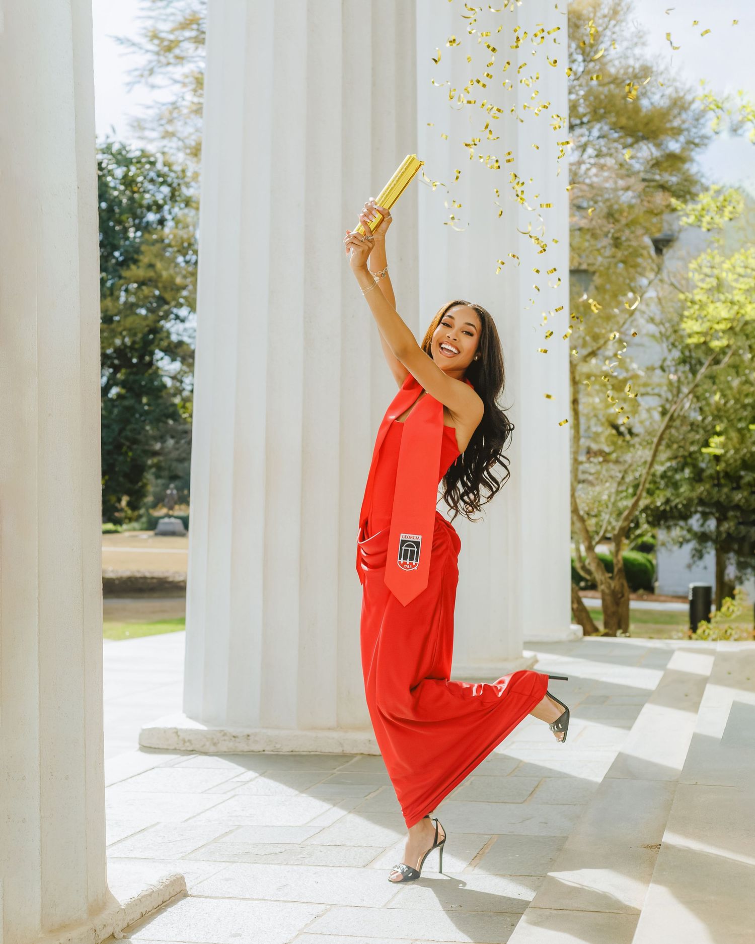 Woman in red jumpsuit celebrating with gold confetti cannon between white columns outdoors.