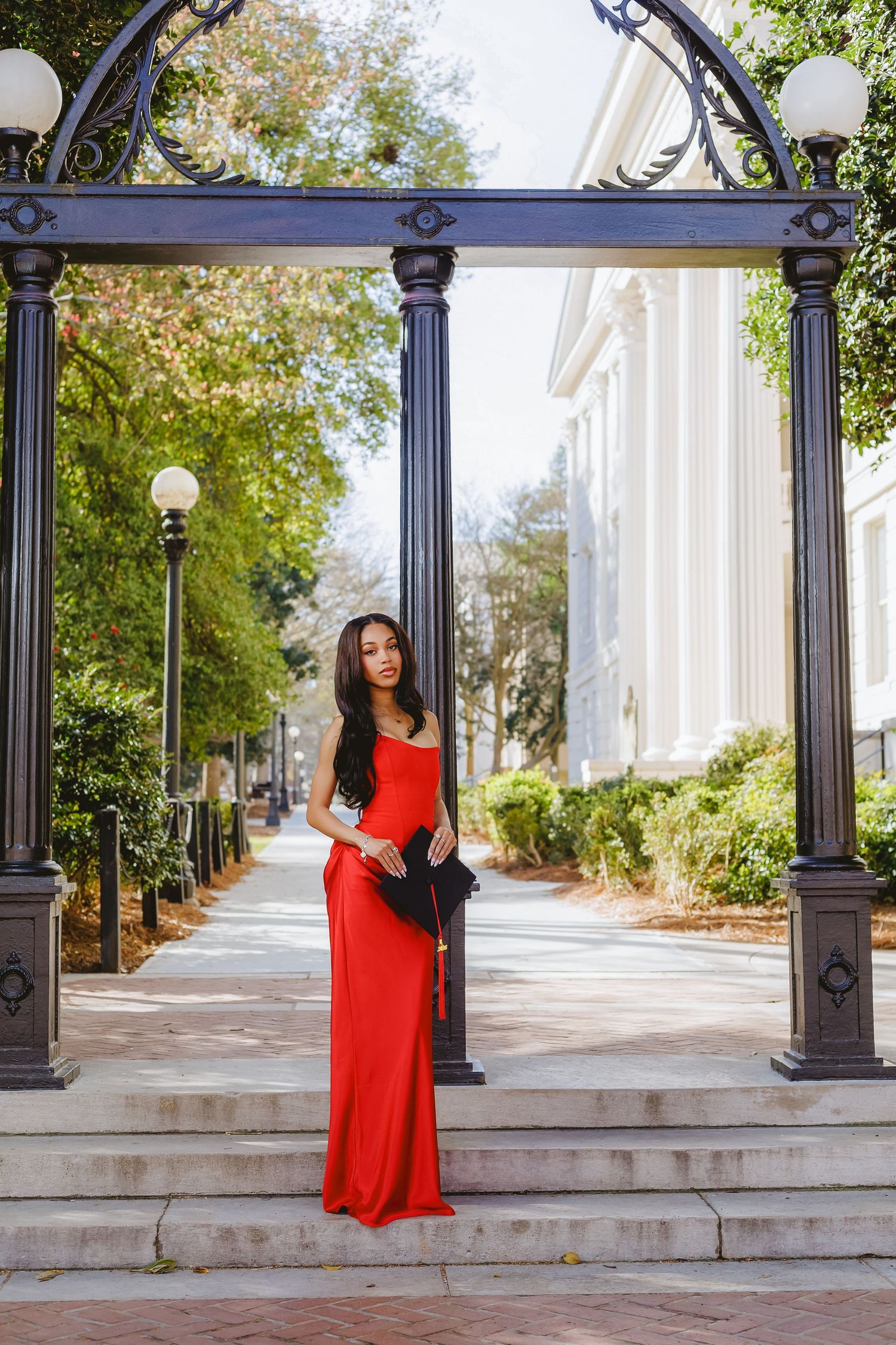 Elegant graduate in red gown standing under ornate arch at university campus entrance.