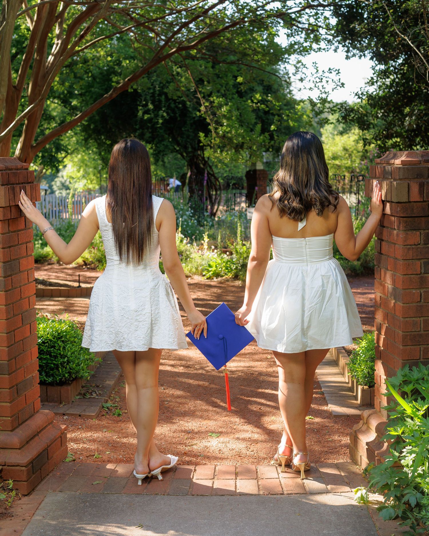 Two women in white dresses walking through brick archway, one holding blue graduation cap.