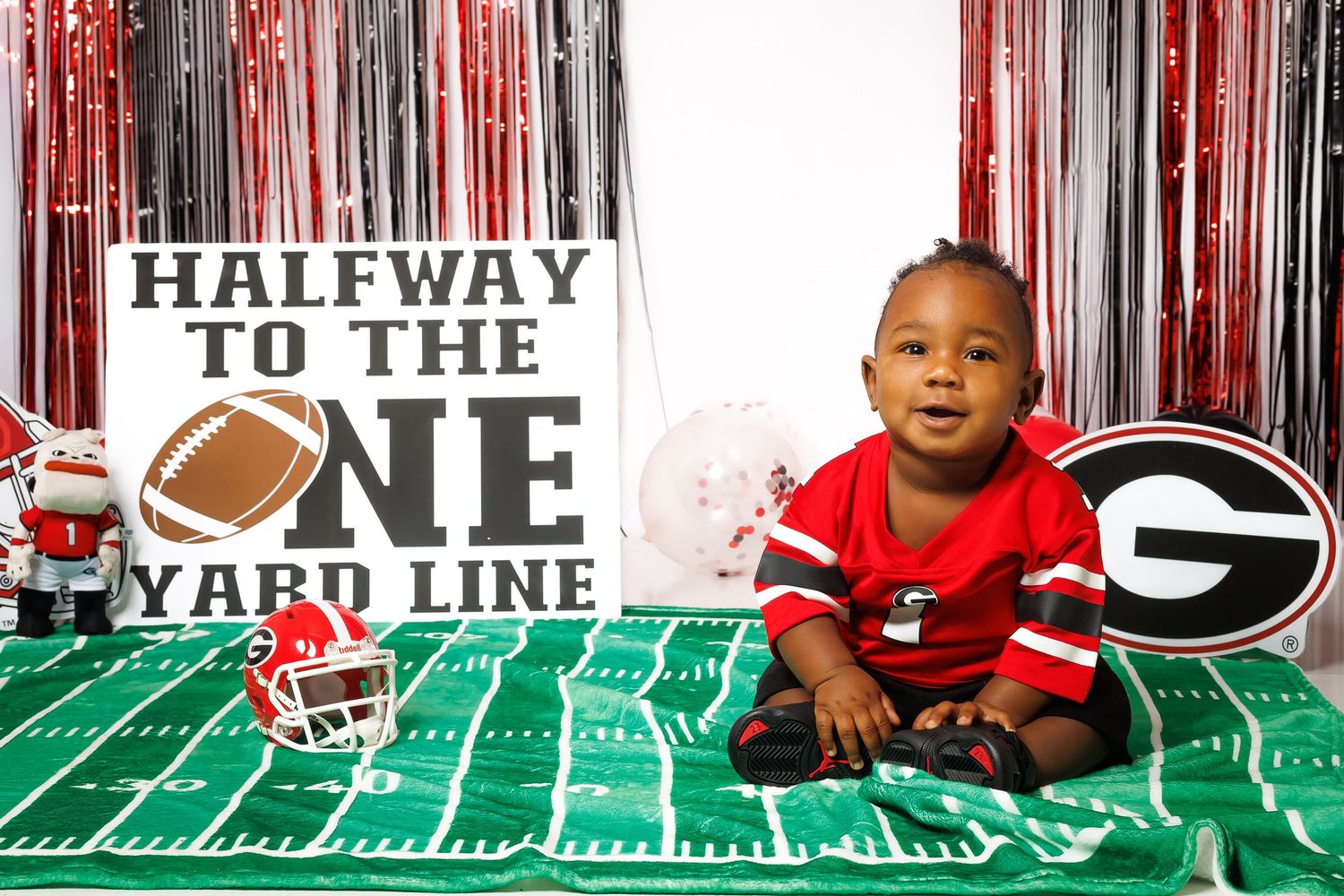 Baby boy in Georgia Bulldogs red jersey sits on football field mat with UGA themed decorations and halfway sign.