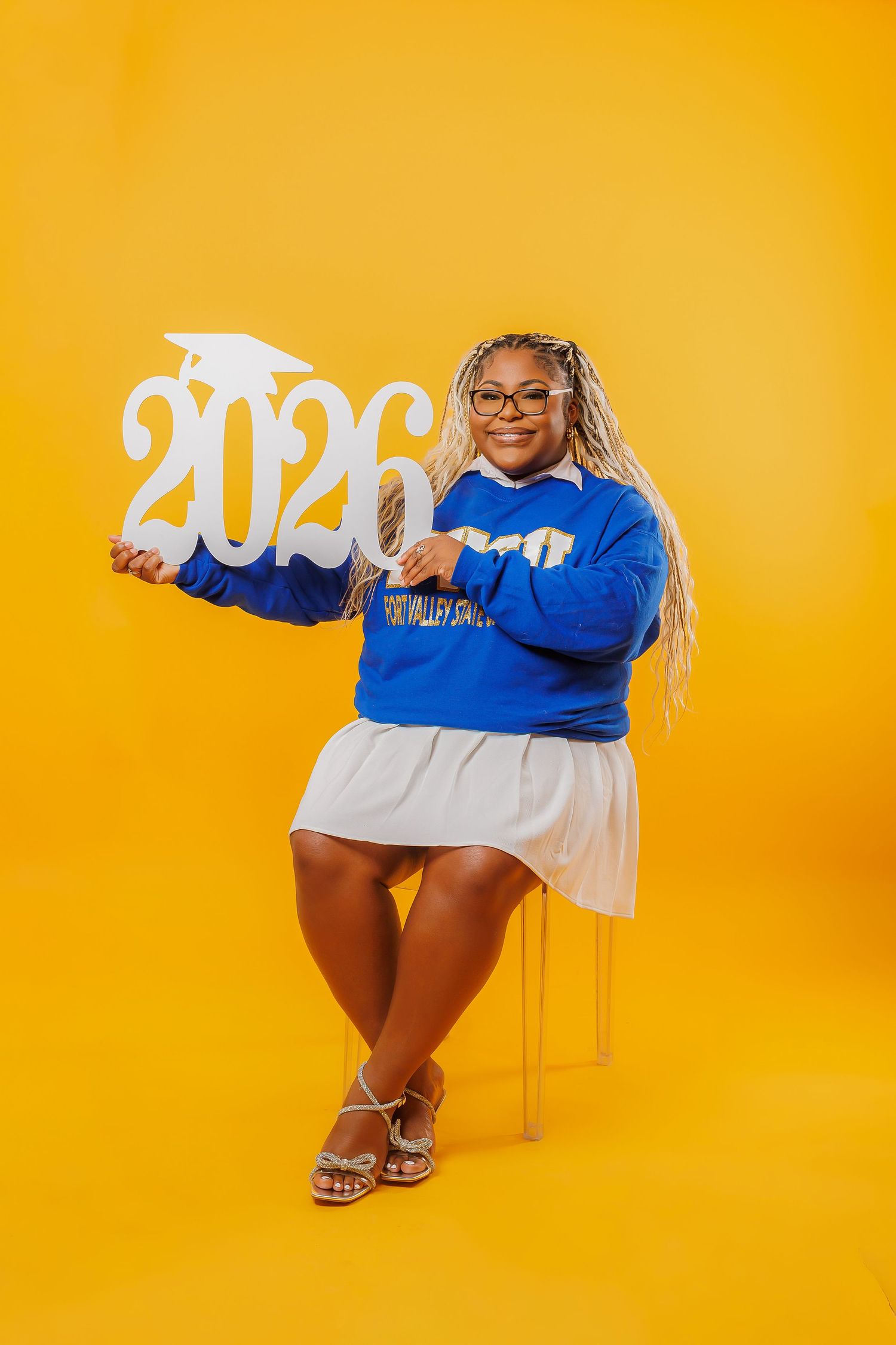 Young woman in blue sweatshirt holds 2026 graduation sign, posing on yellow background for senior photos.