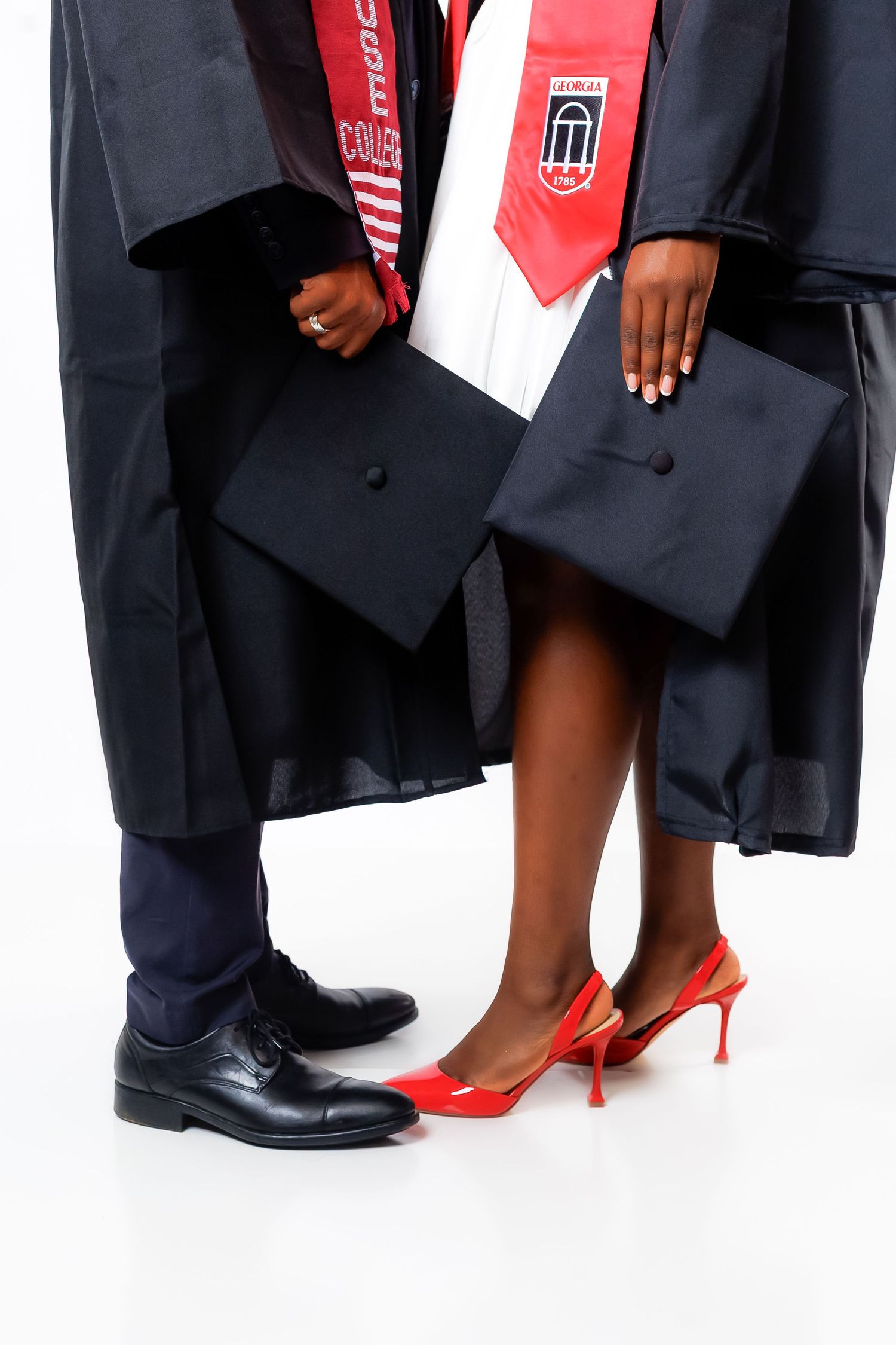 Close-up of two graduates in black robes holding caps, one wearing red heels, white background.