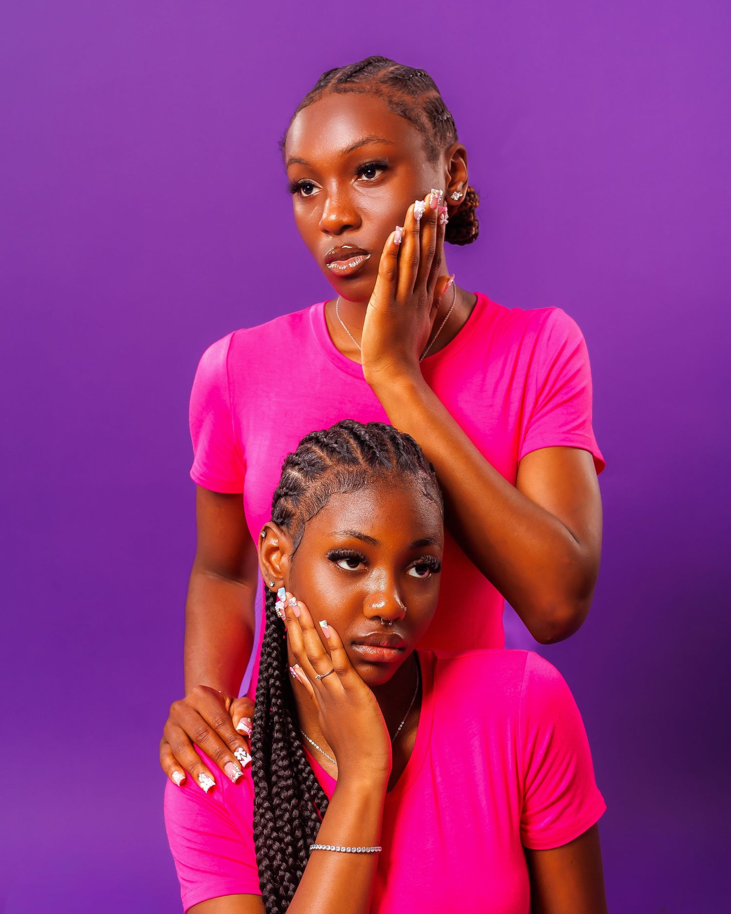 Two young Black women in matching pink t-shirts pose together against a vibrant purple background.
