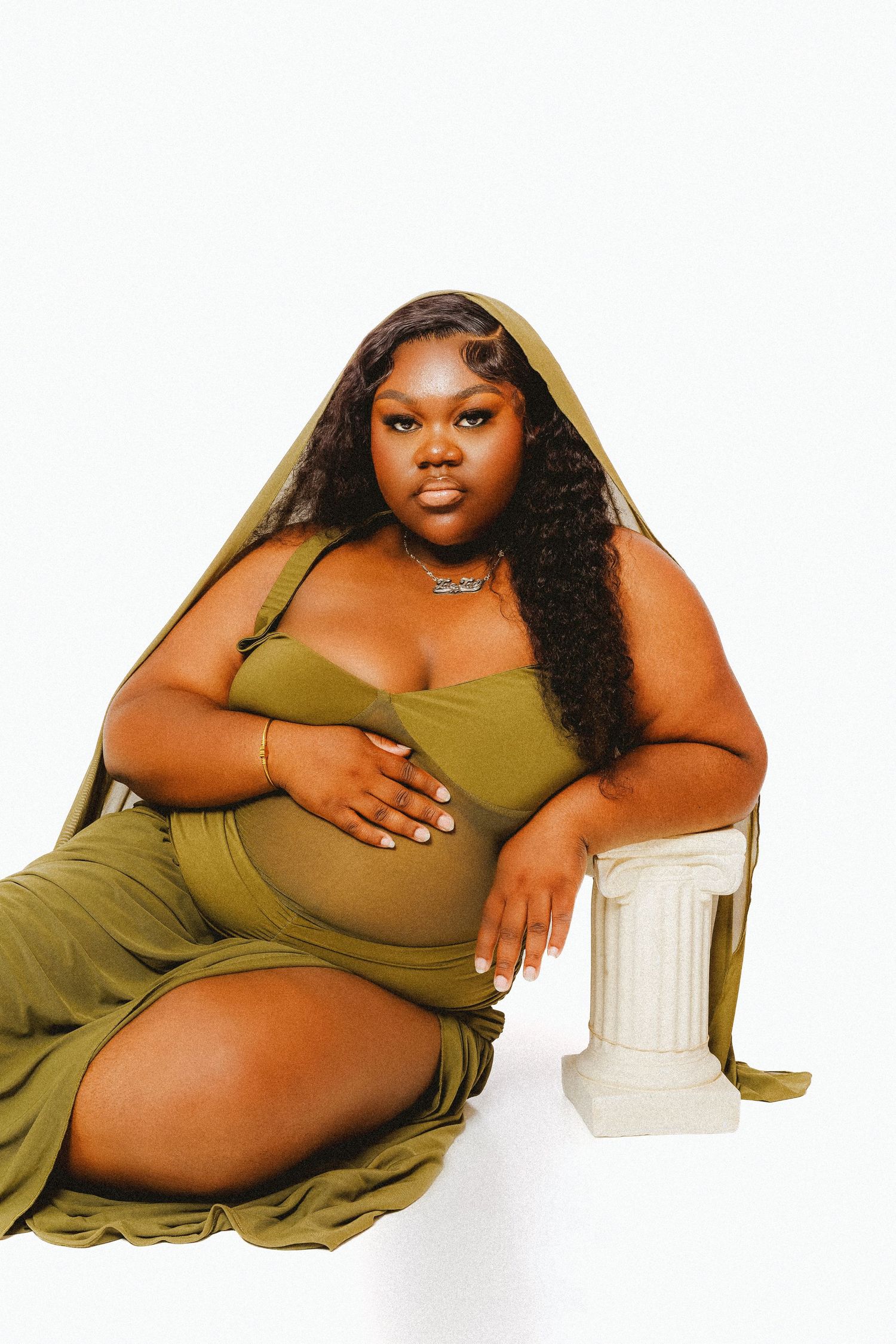 A plus-size woman poses elegantly in an olive green maternity dress beside a white pedestal column.