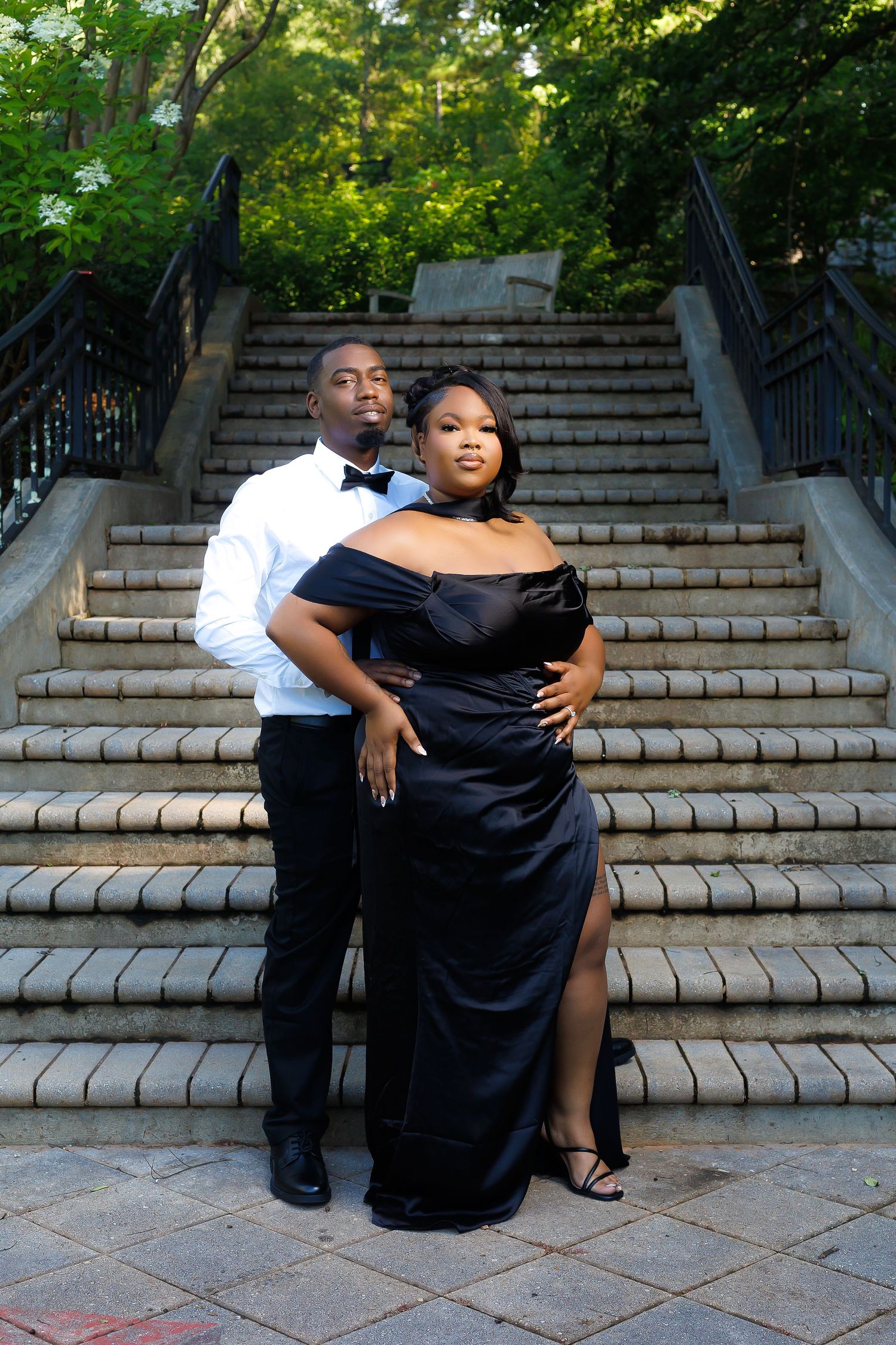 Couple in elegant black formal wear posing on stone steps surrounded by lush greenery.