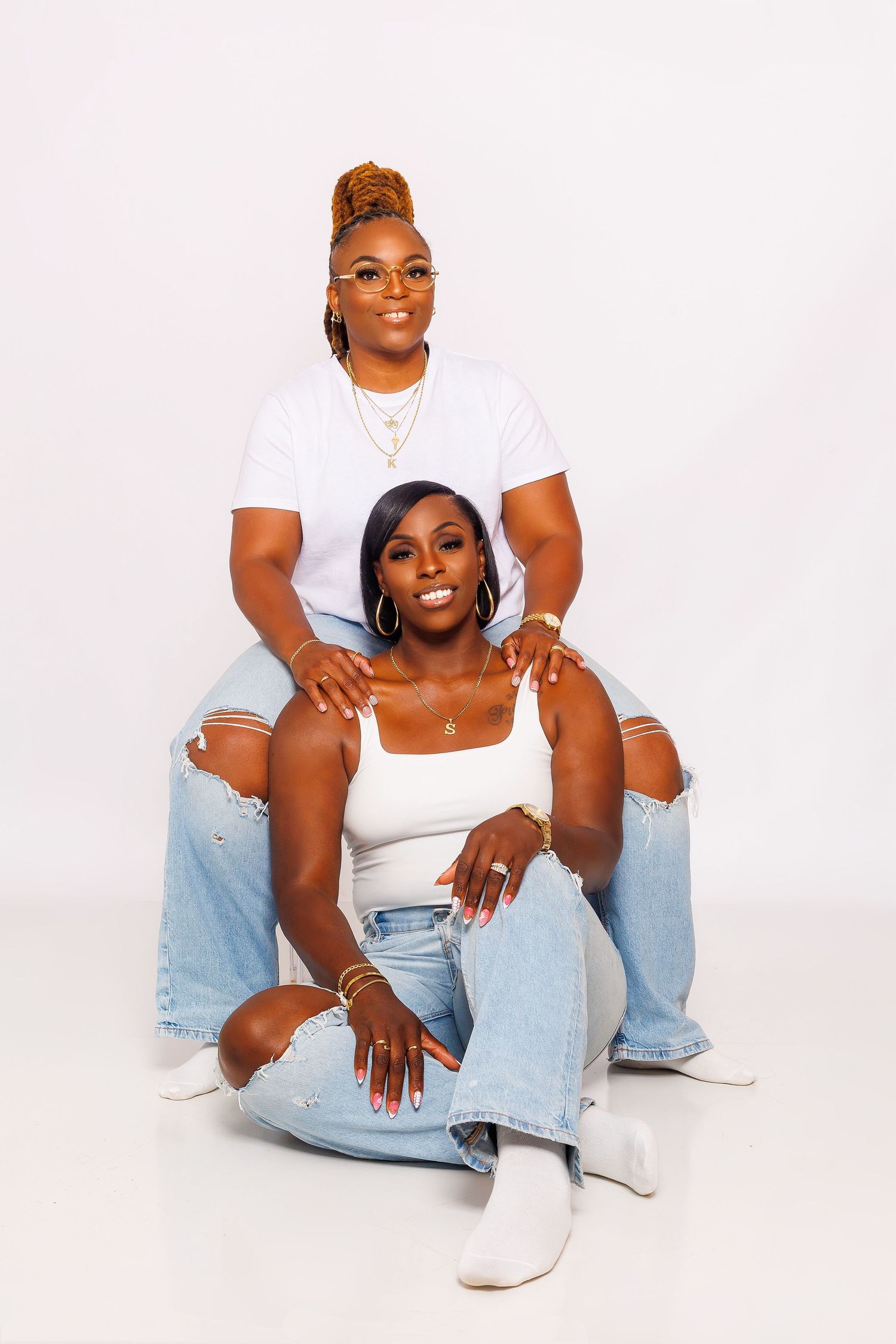 Two women in matching white tops and ripped jeans posing together on white background.