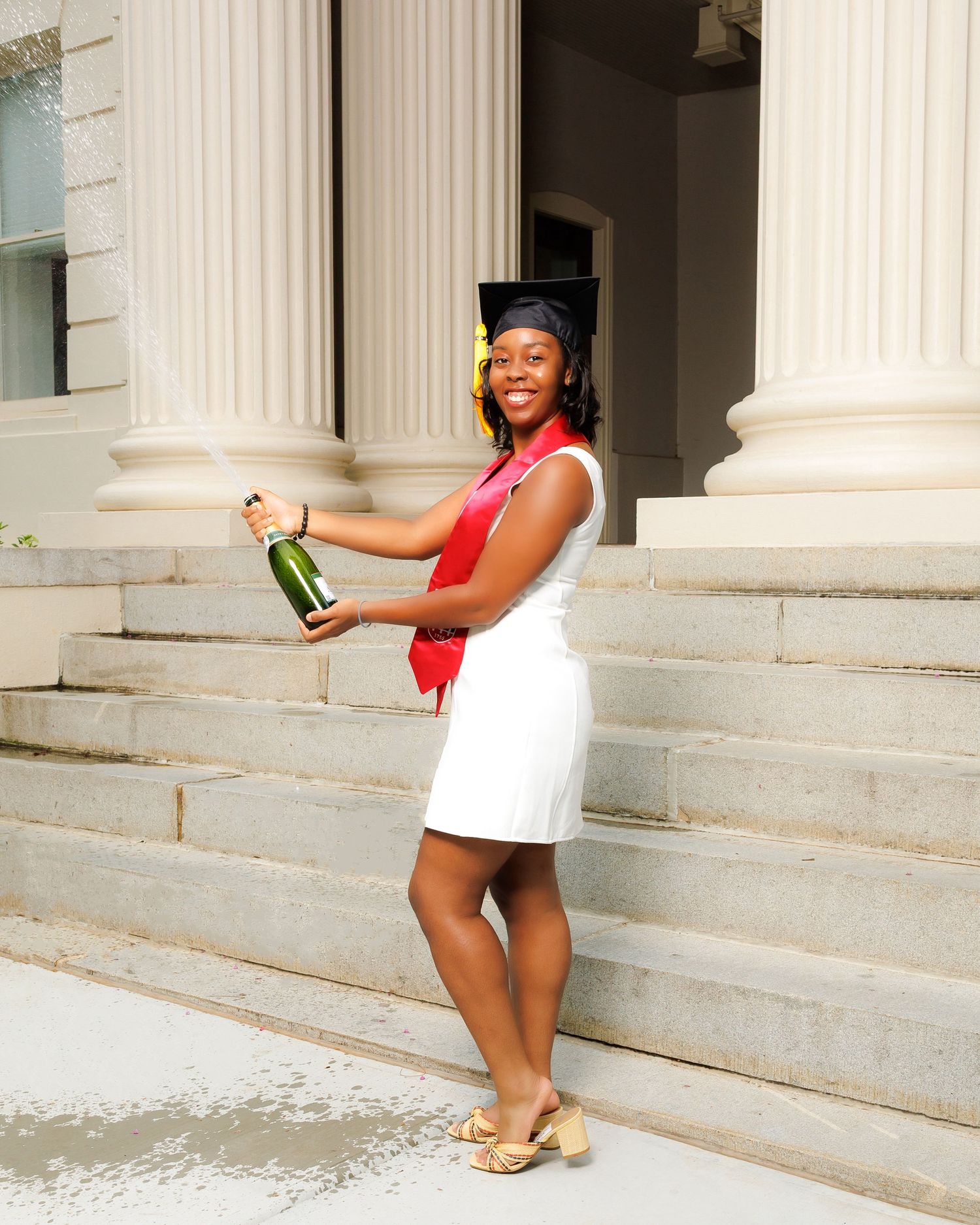 Smiling graduate in white dress and cap popping a champagne bottle on university building steps.