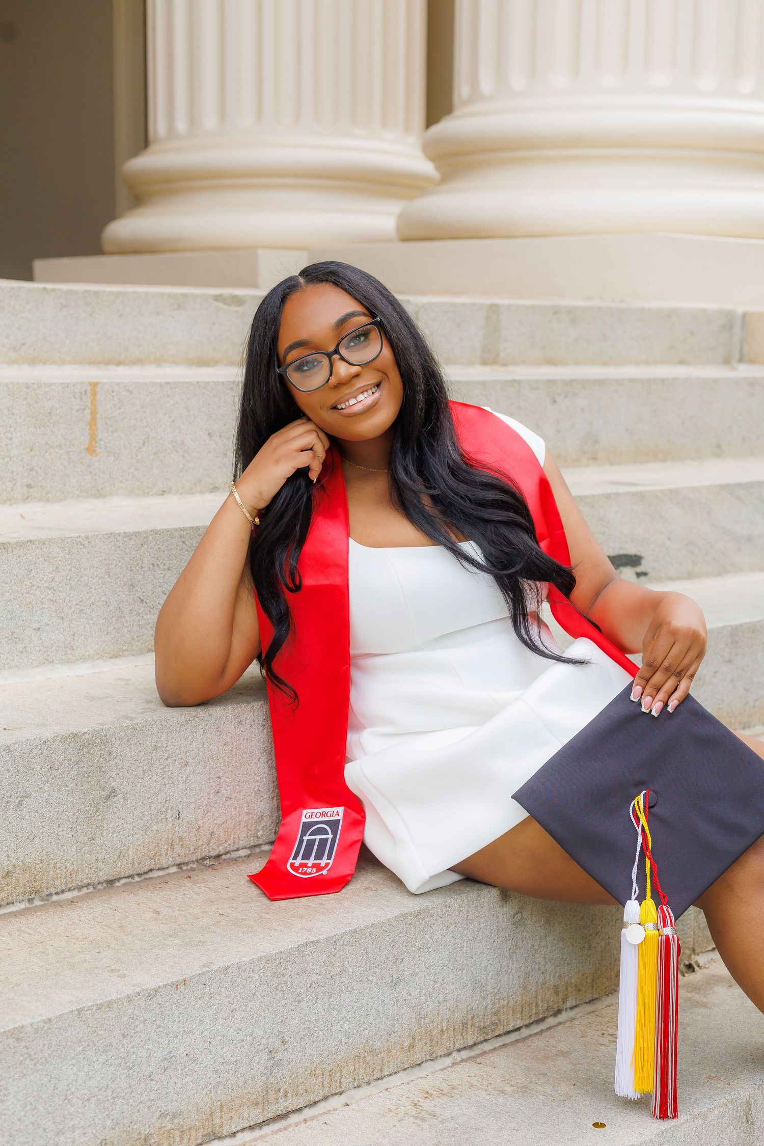 Smiling graduate sitting on marble steps holding cap with red graduation stole and glasses.
