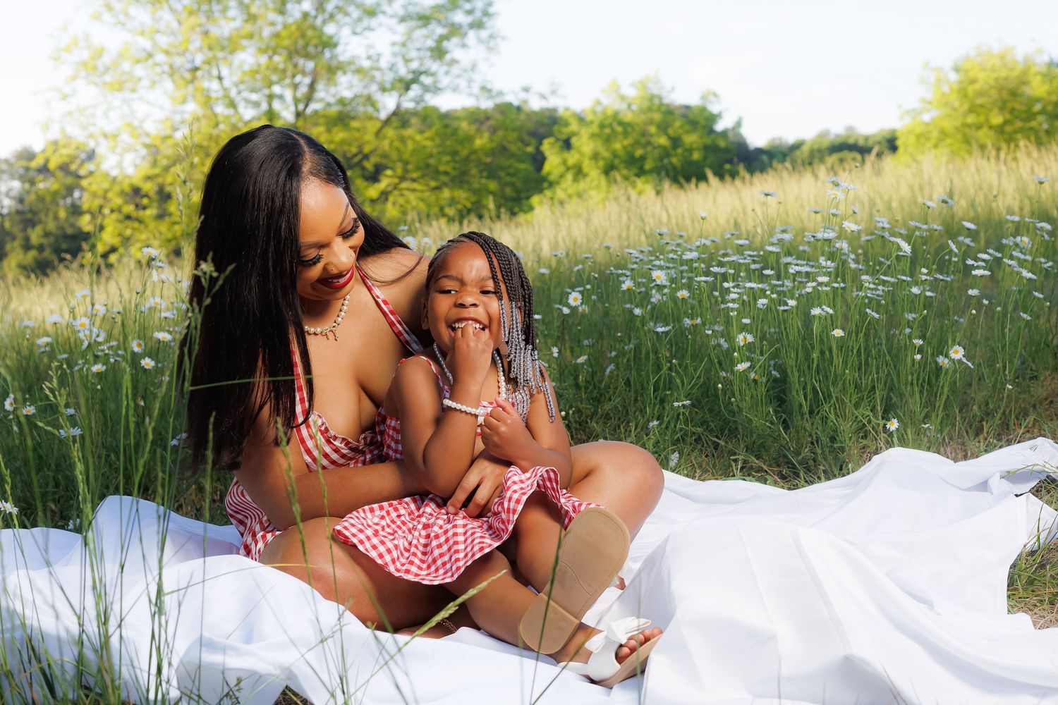 A mother and young daughter share a joyful moment on a white blanket in a sunny wildflower meadow.