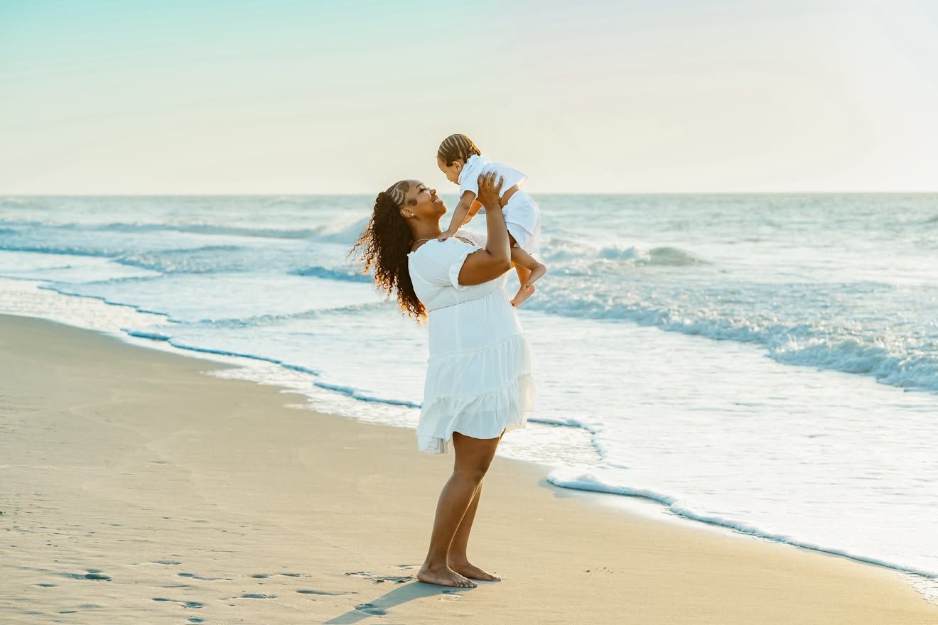 A mother in a white dress joyfully lifts her baby on a sunny beach with ocean waves in the background.