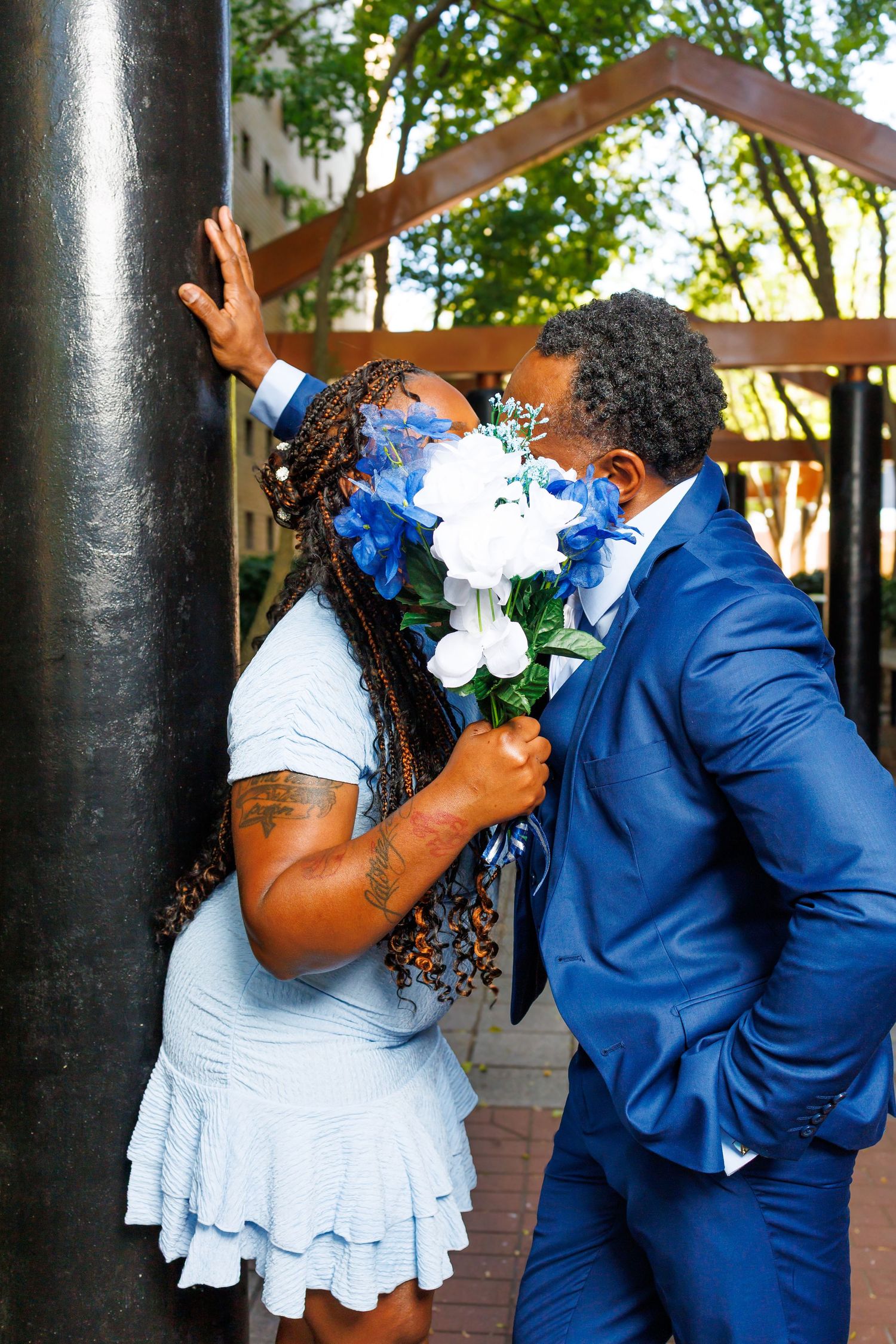 Couple sharing a kiss hidden behind blue and white wedding bouquet near a dark pillar outdoors.