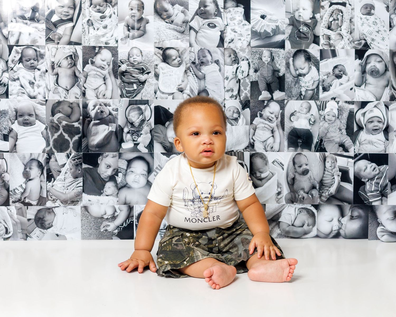 Baby in Moncler onesie and camo pants sits before wall covered in black and white baby photos.