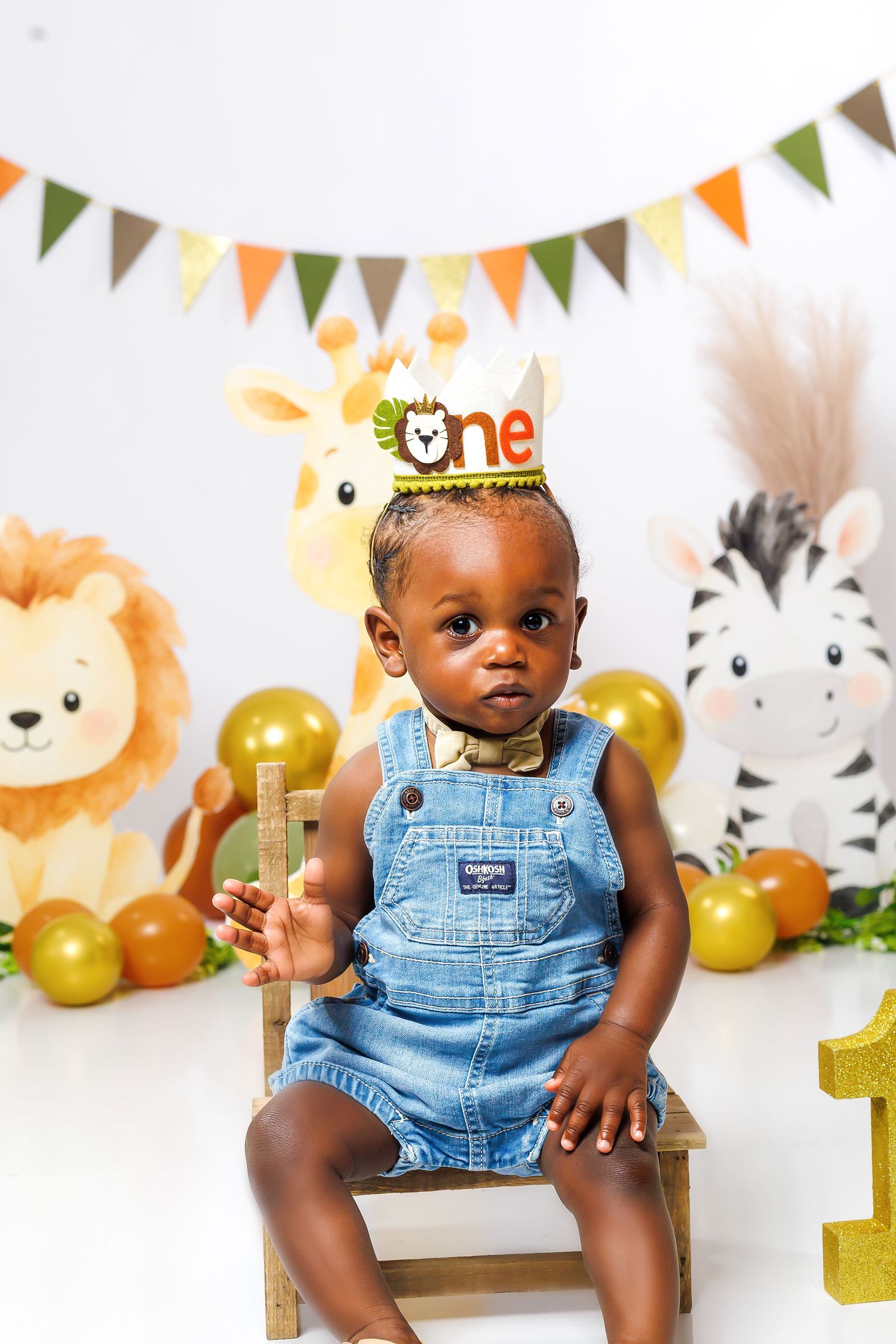 Baby boy in denim overalls wearing safari birthday crown seated during first birthday photoshoot.