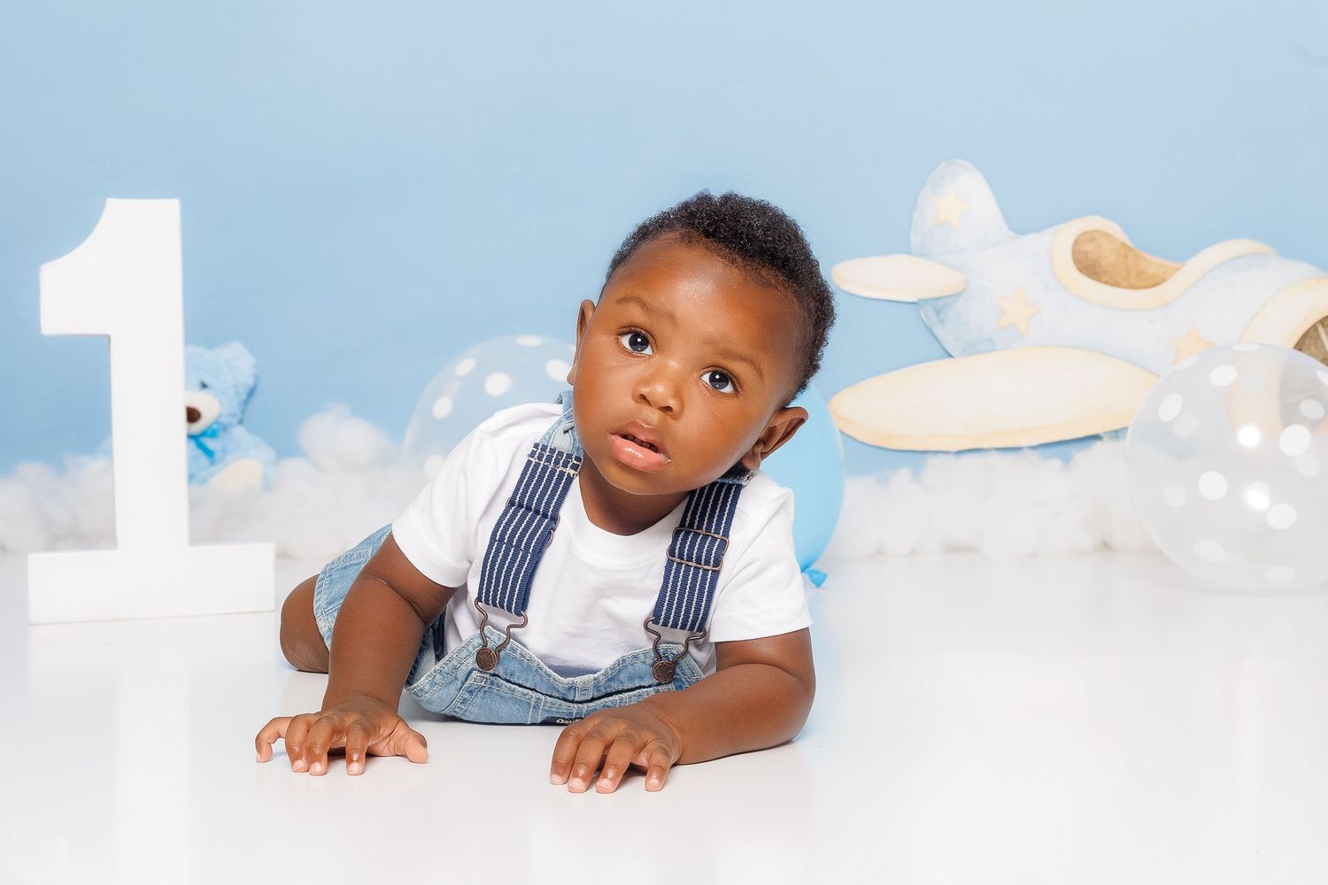 Adorable baby in denim overalls posing near number one decoration for first birthday photo.
