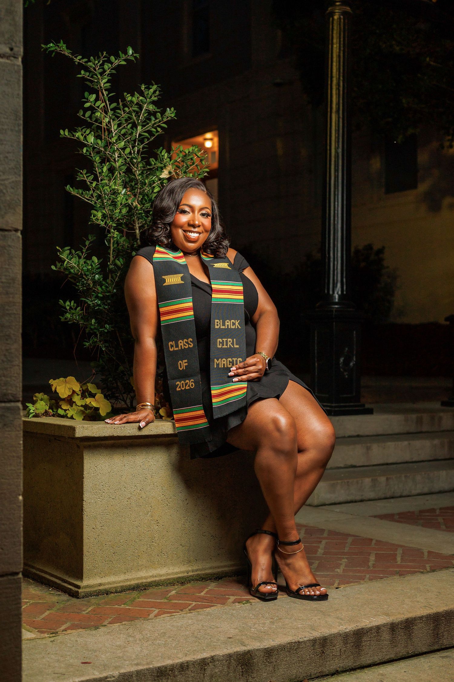 Smiling graduate wearing Kente cloth stole reading Black Girl Magic Class of 2020 seated outdoors.