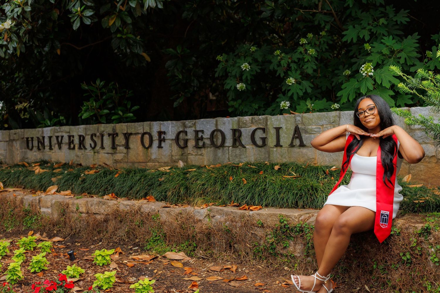 Graduate in white dress and red stole posing beside University of Georgia stone sign outdoors.