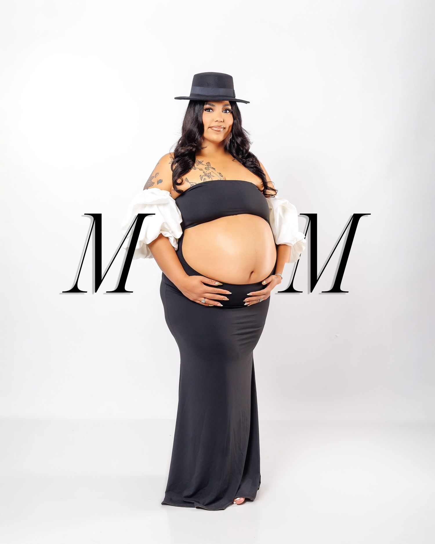 Pregnant woman in black maxi skirt and bandeau top with white jacket and hat poses for maternity photo shoot.
