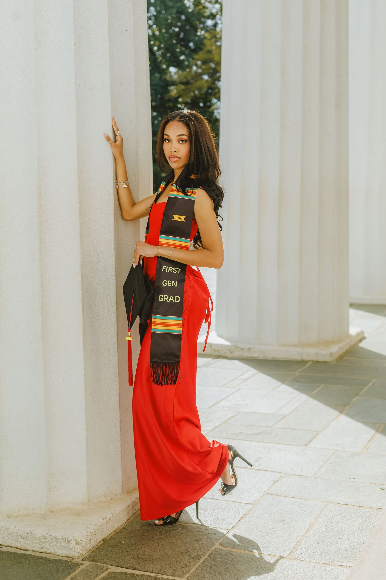 Young woman in red gown holding First Gen Grad stole and cap, posing between white columns for graduation photos.