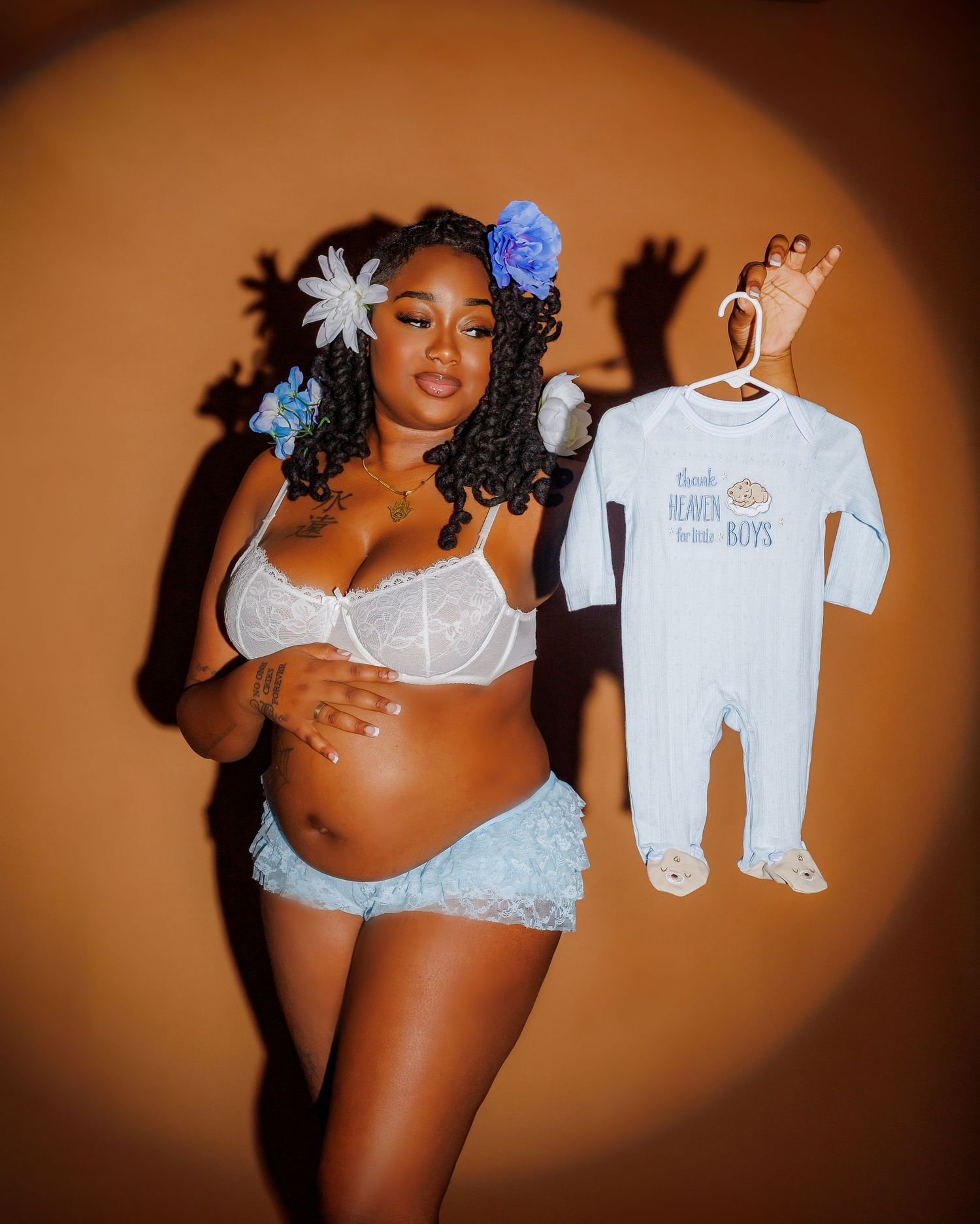 Pregnant Black woman holds baby boy onesie wearing white lace lingerie with blue floral hair accessories.