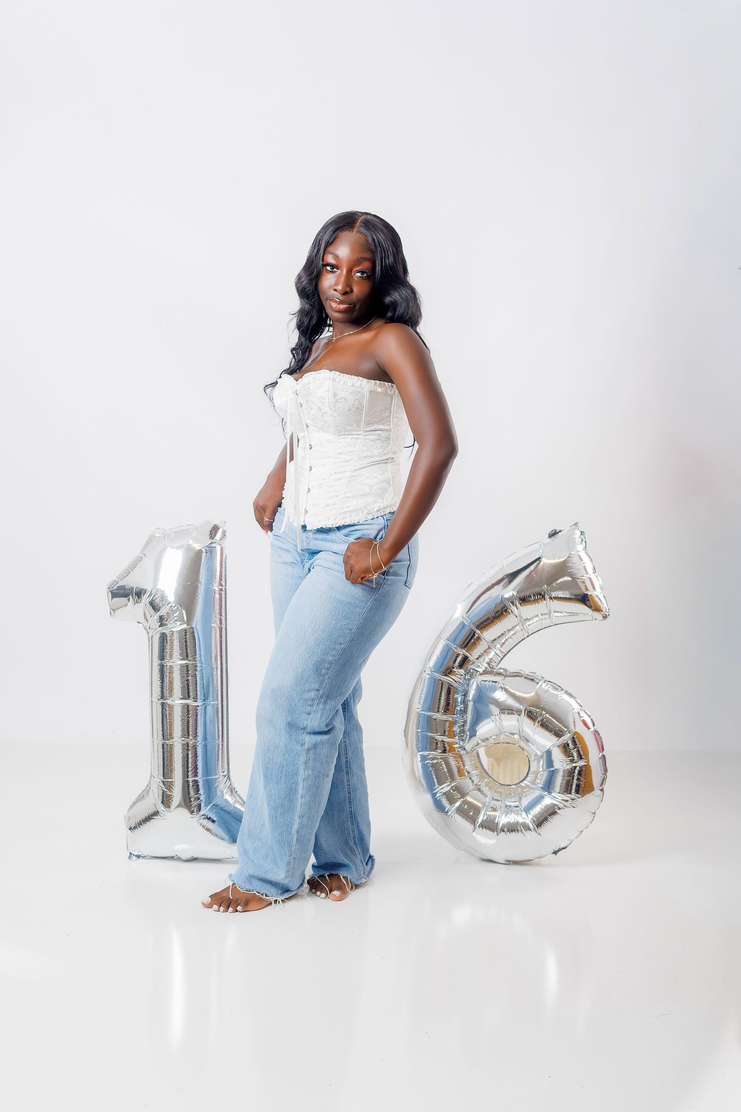 Young Black woman poses with silver number 16 balloons wearing white corset top and flared jeans on white background.