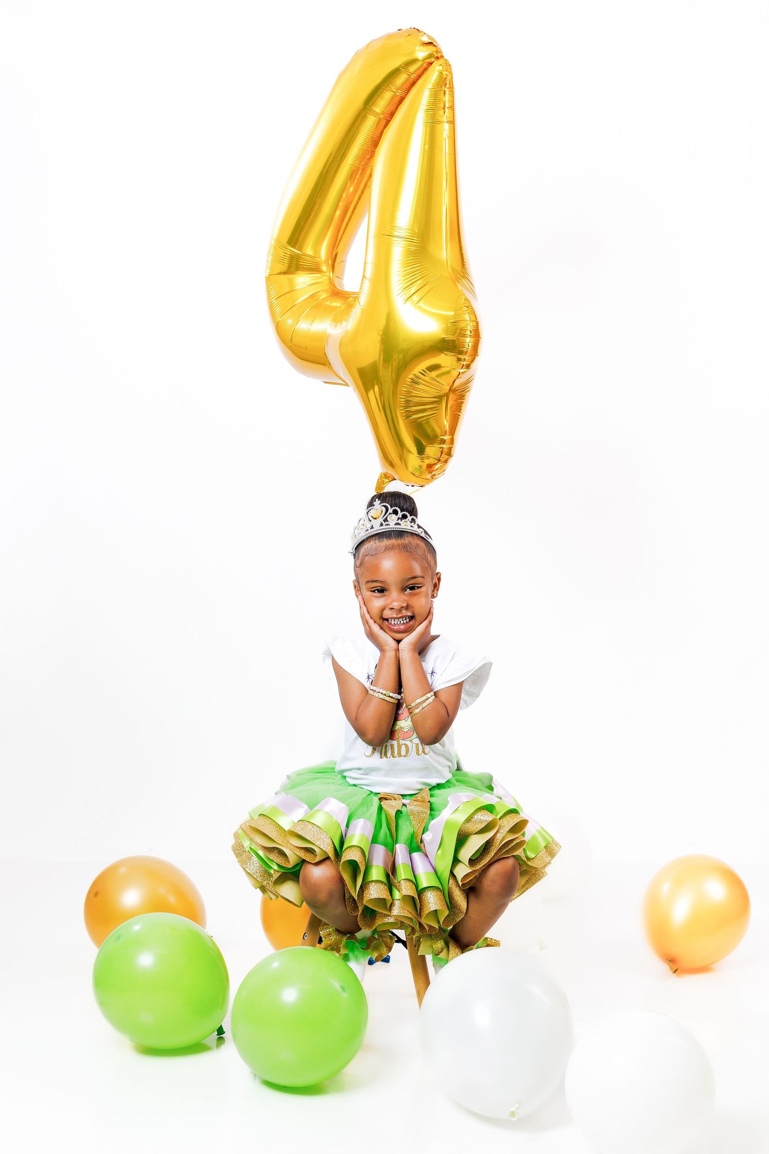 Young girl in princess dress sits cross-legged under large gold number 4 balloon with colorful balloons around her.