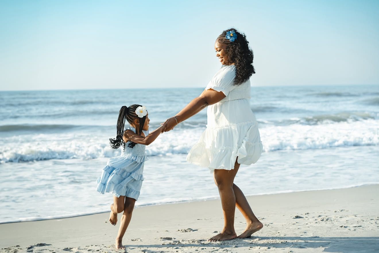 A mother and daughter in white dresses hold hands and dance joyfully on a sunny beach by the ocean.