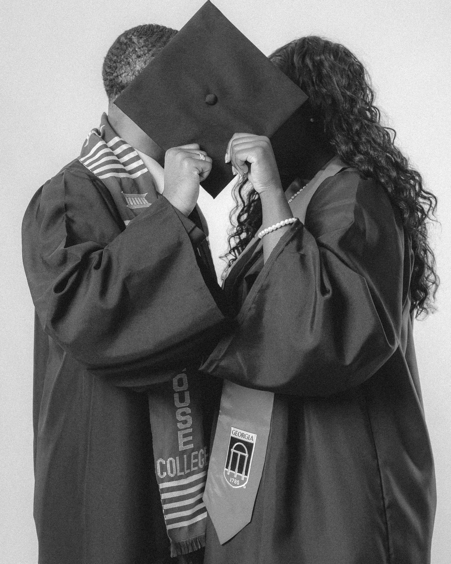 Black and white photo of two graduates hiding faces behind a shared graduation cap kissing.