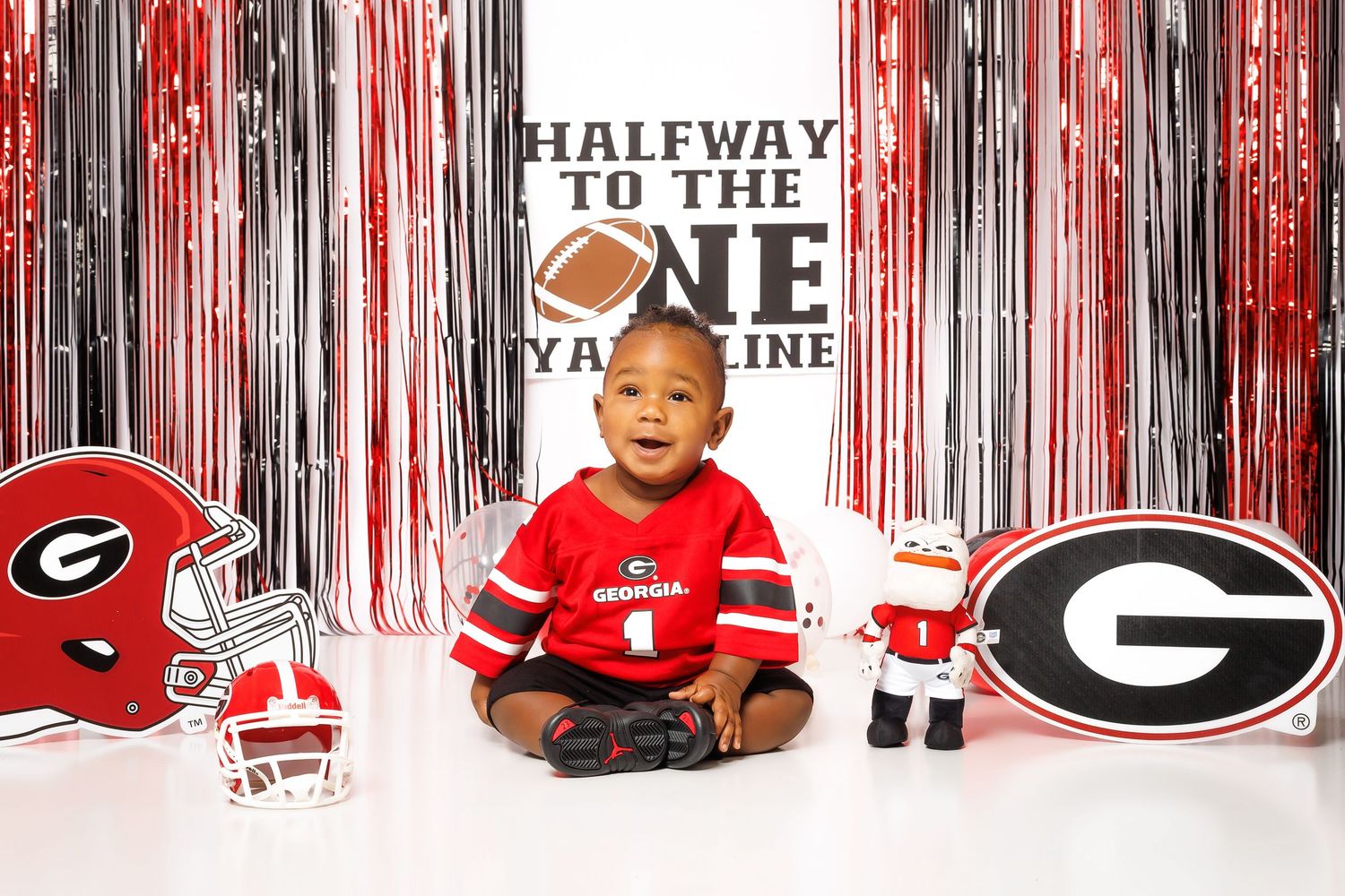 Smiling baby in Georgia Bulldogs jersey sits between UGA helmets and mascot toy with themed backdrop.