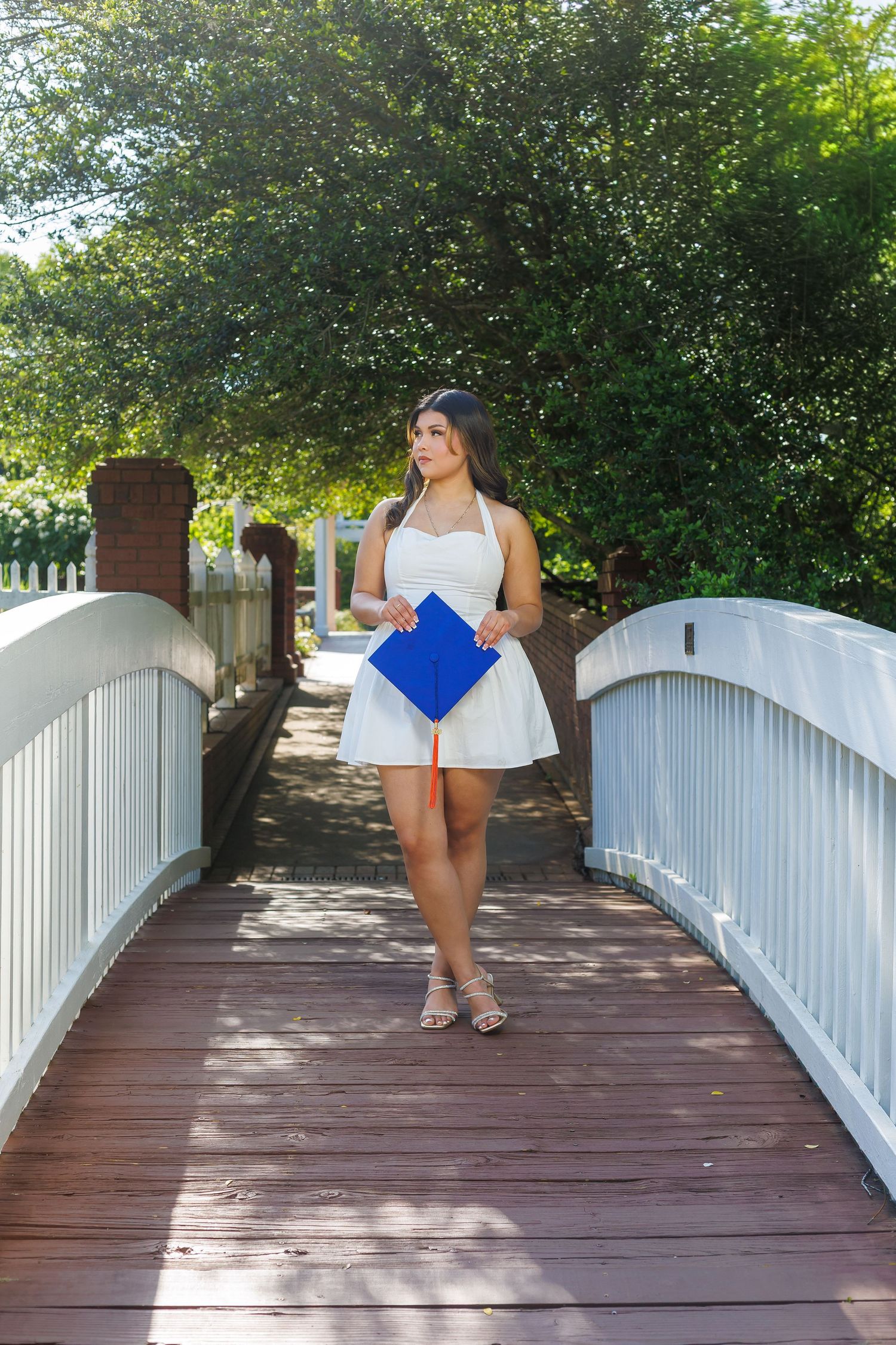 Young woman in white dress holding blue graduation cap while walking across wooden bridge outdoors.