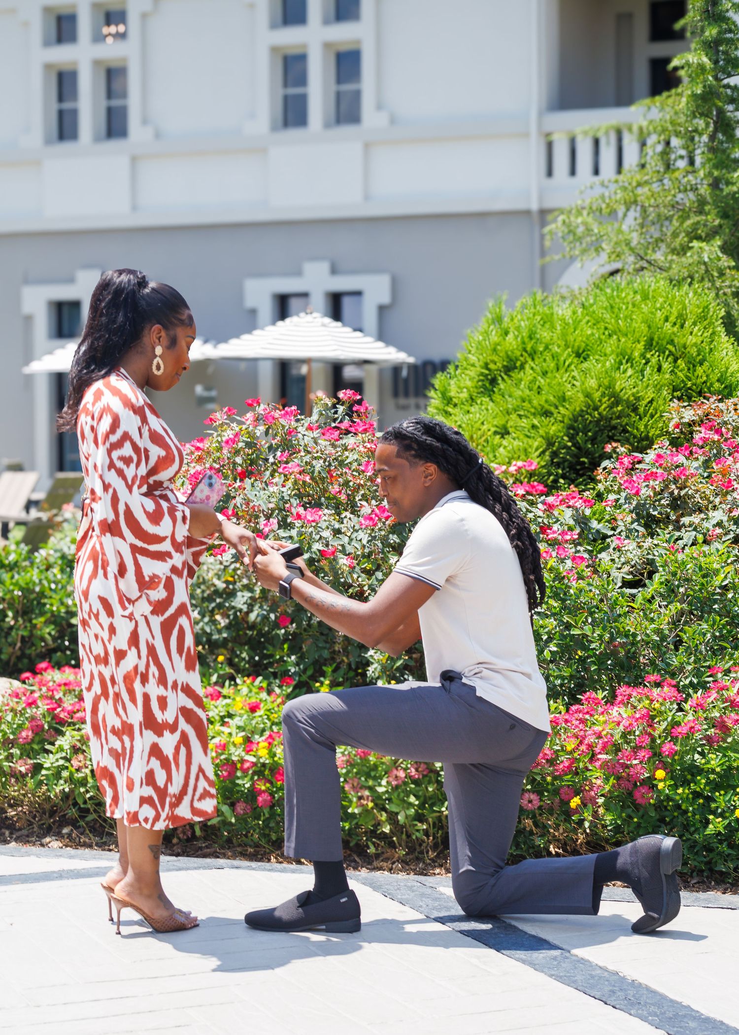 Man presenting engagement ring while kneeling before woman in red dress in a garden setting.