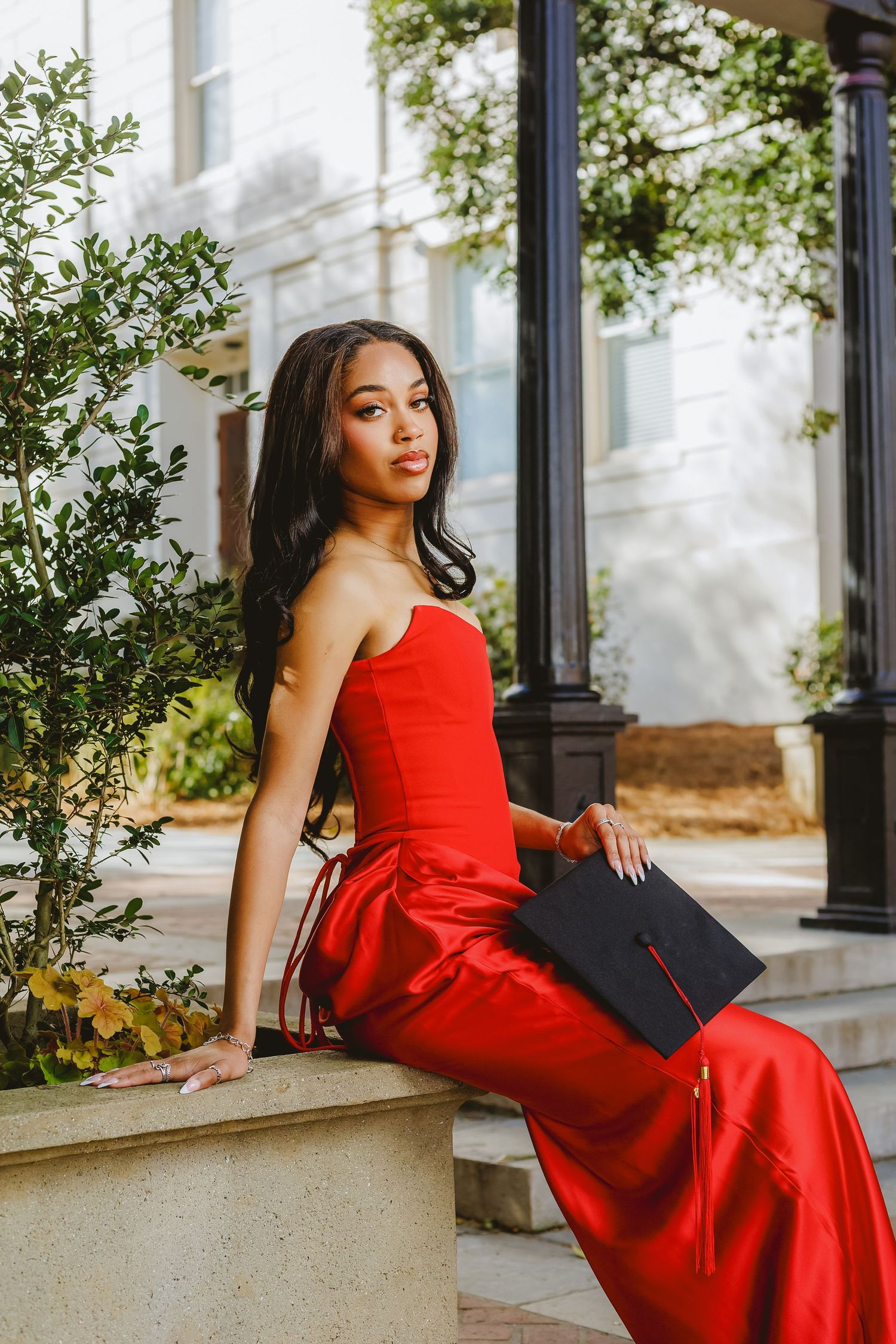 Young woman in elegant red strapless gown holding graduation cap while seated outdoors.