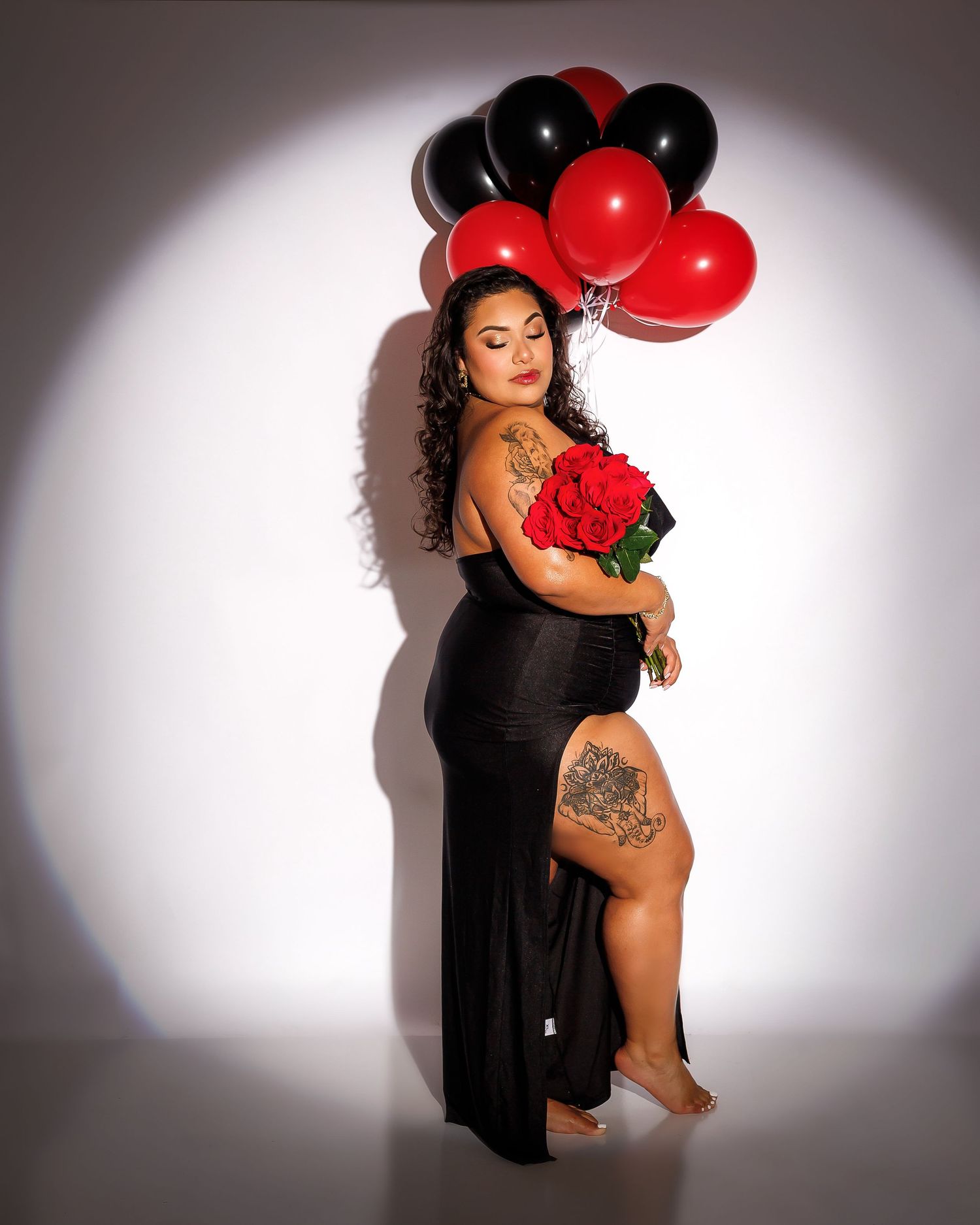 Curvy woman in black dress holds red roses and red and black balloons in dramatic spotlight.