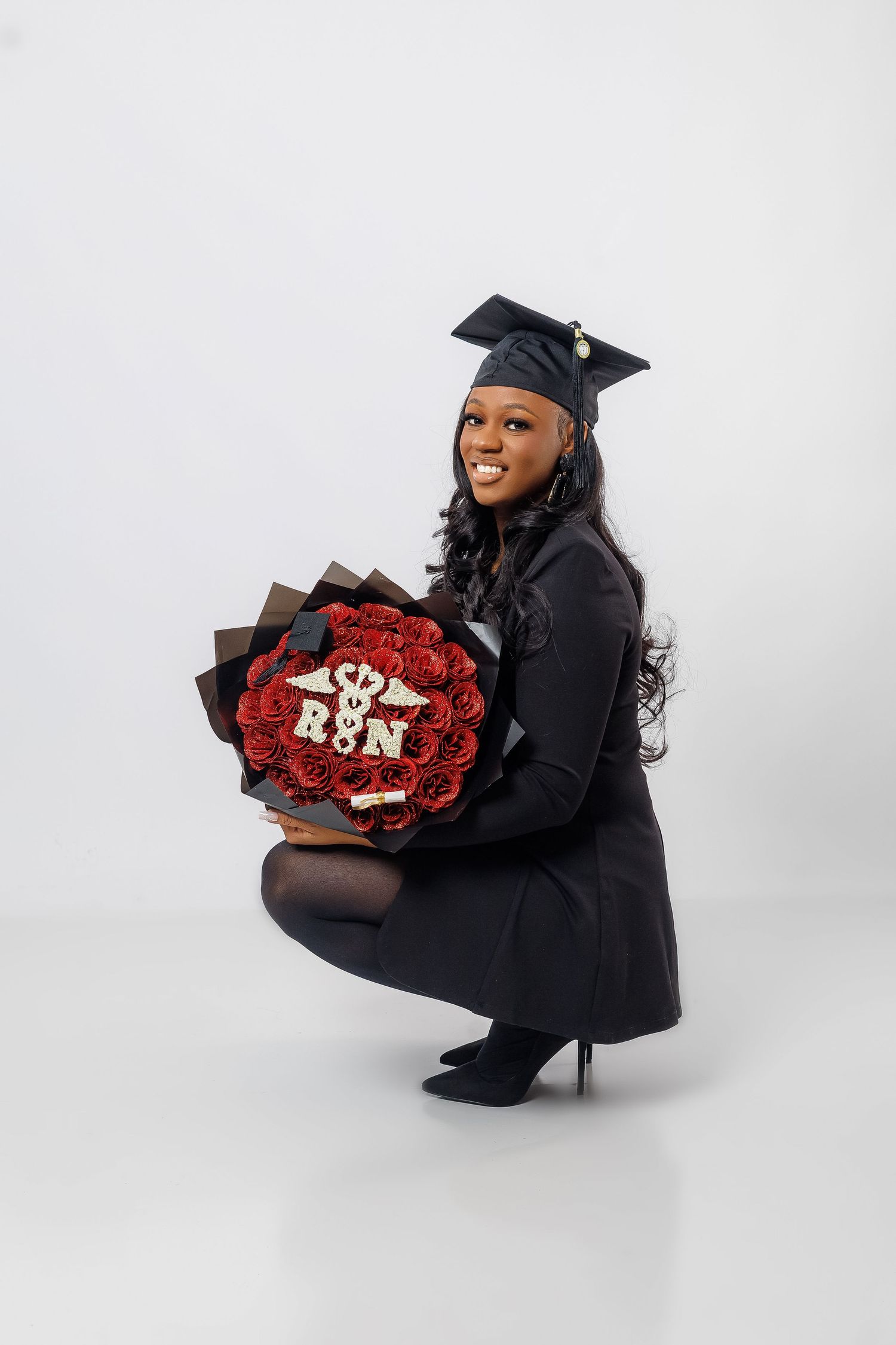 Smiling nursing graduate in black cap and gown crouching with red rose bouquet featuring RN emblem.