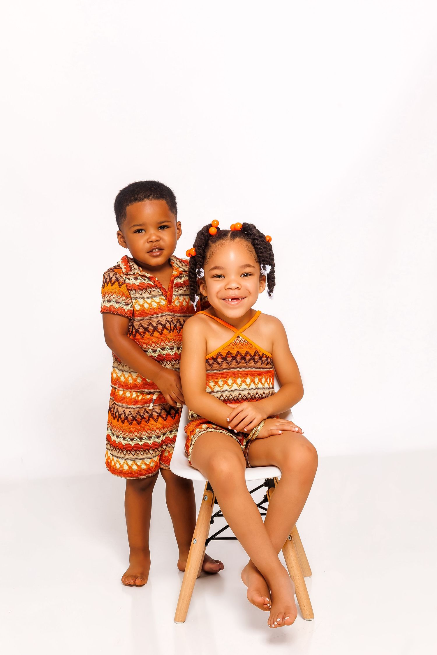 Two young siblings in matching orange patterned outfits pose together on white background, girl seated on stool.