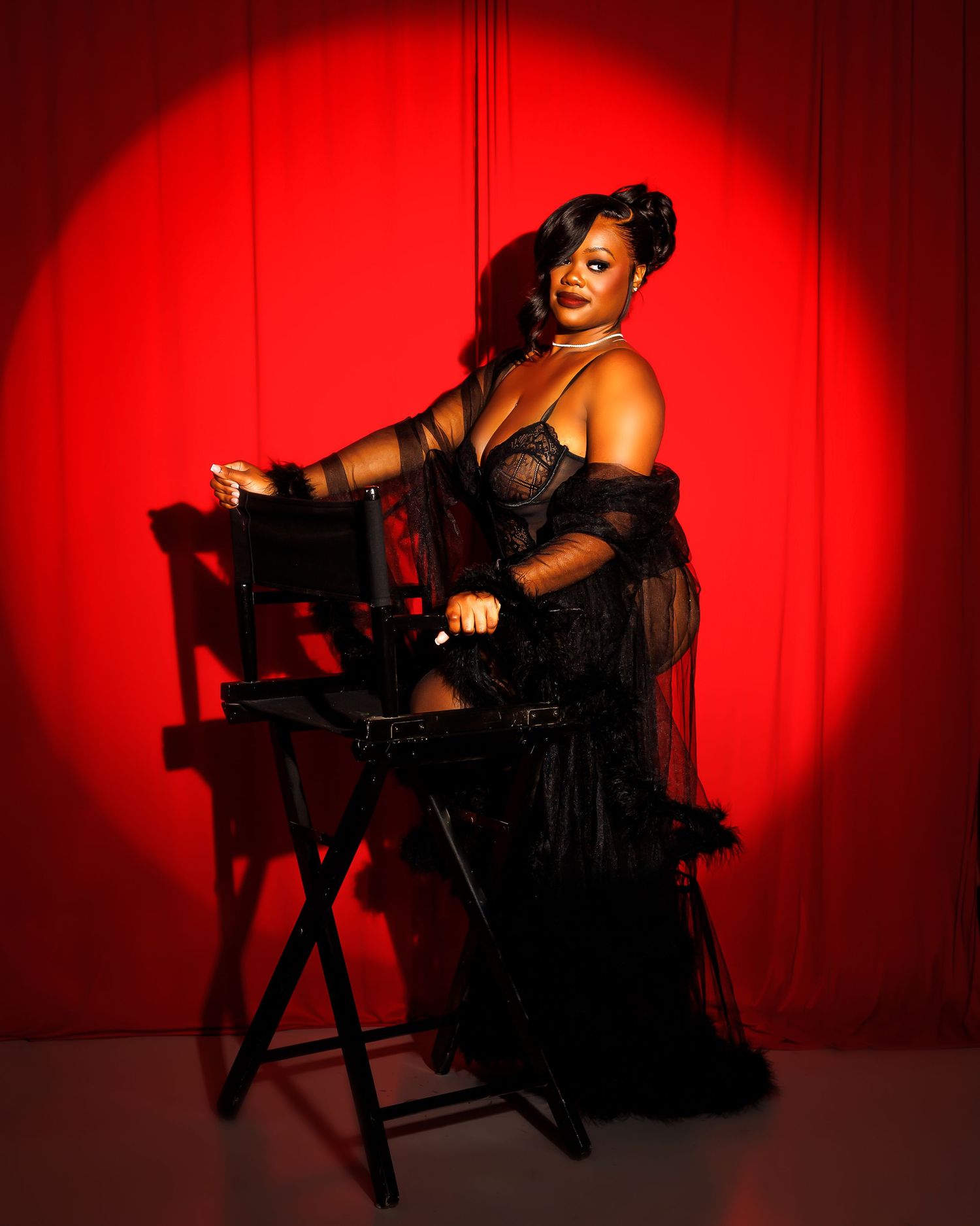 Woman in black sheer gown poses elegantly on director's chair against dramatic red spotlight background.