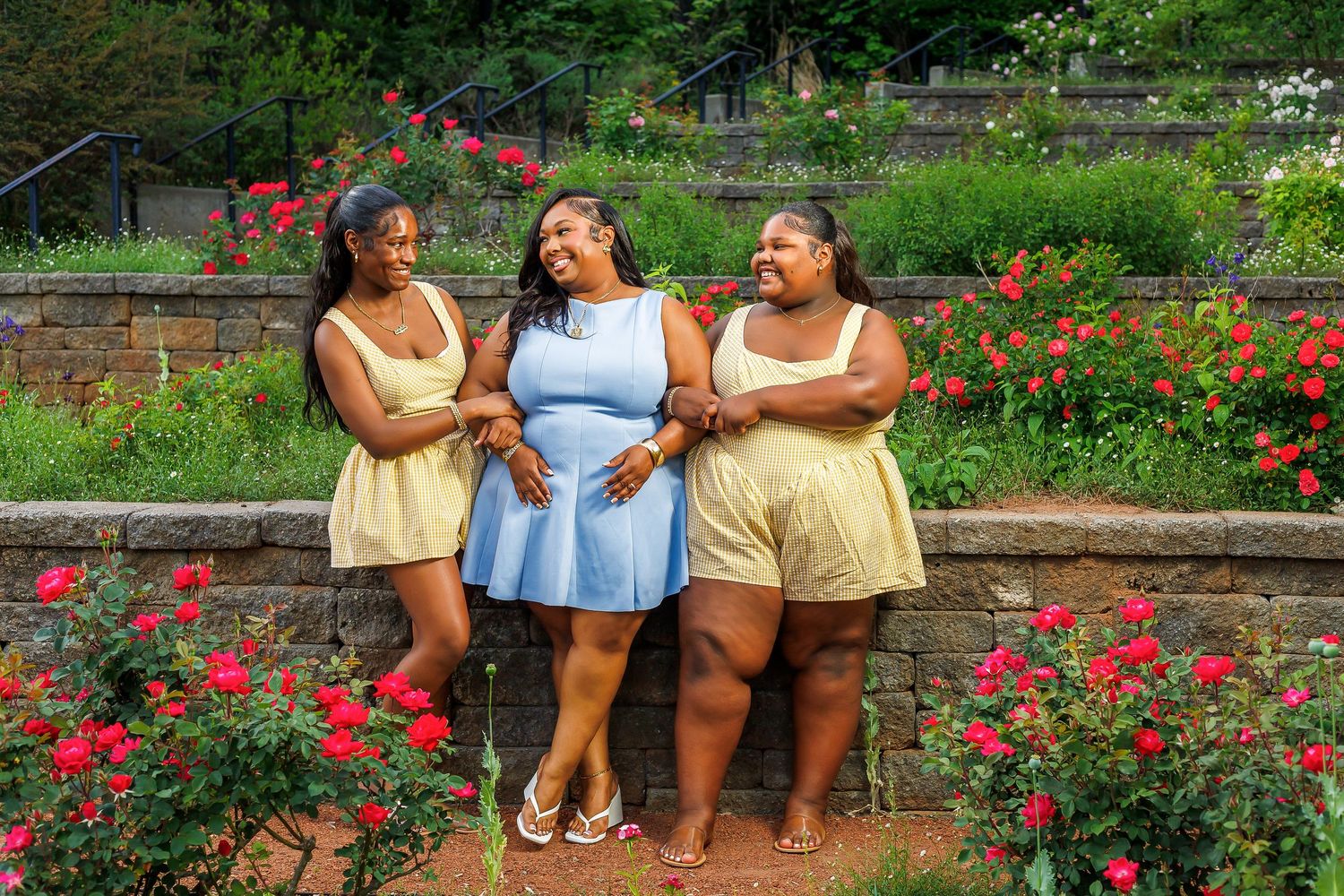Three women in colorful summer outfits smiling together in a garden with red roses and stone wall.